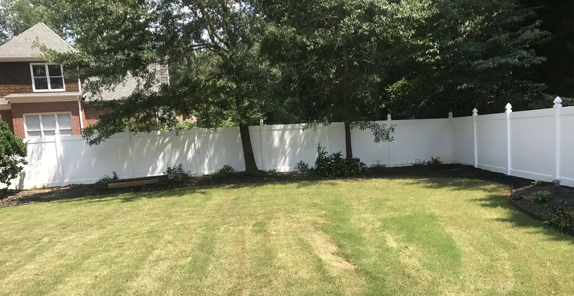 Sunny backyard with striped grass, white fence, trees, and a house partly visible on the left