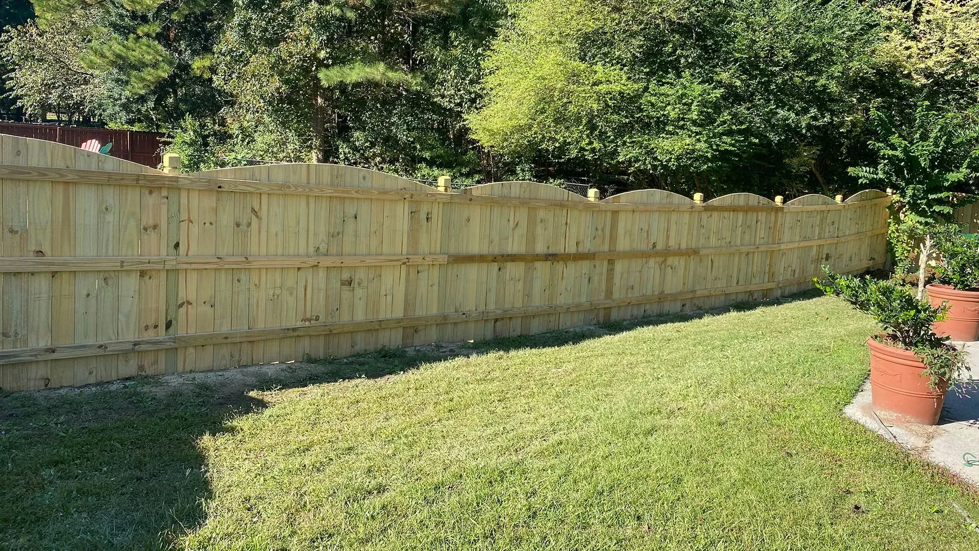 A grassy backyard with a wooden fence, trees beyond it, and potted plants near the patio.
