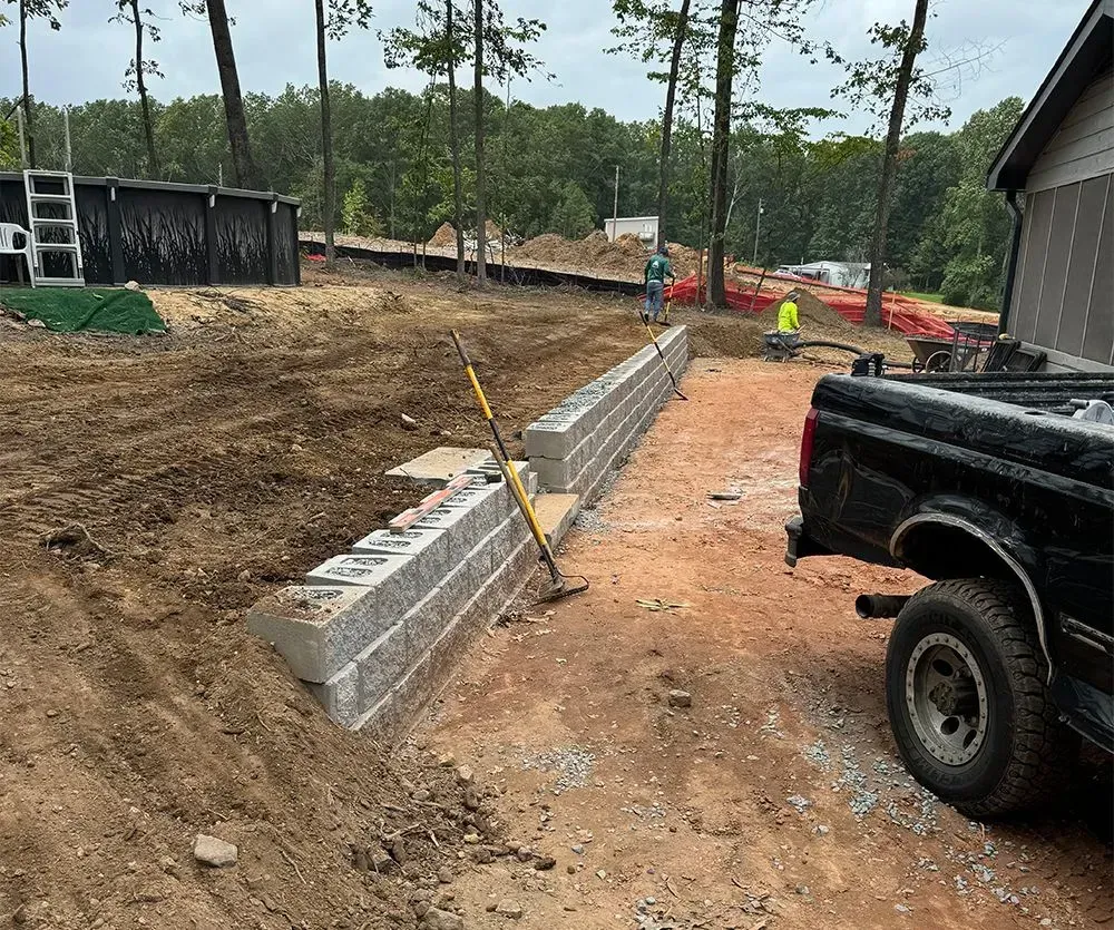 Construction site with a long concrete wall, dirt lot, trees, and a black pickup truck in the foreground.