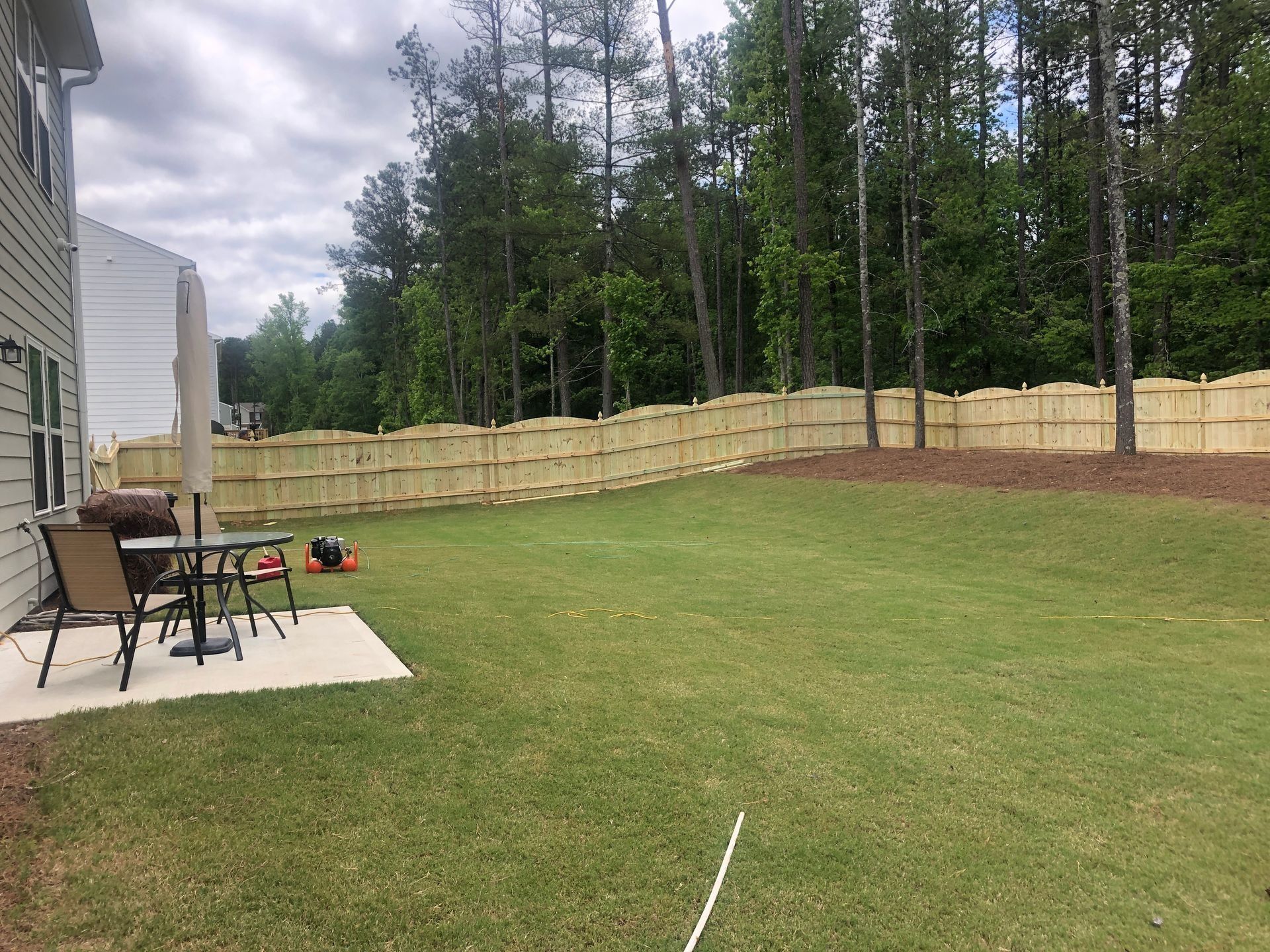 Backyard lawn with patio furniture, beige fence, and trees under a cloudy sky