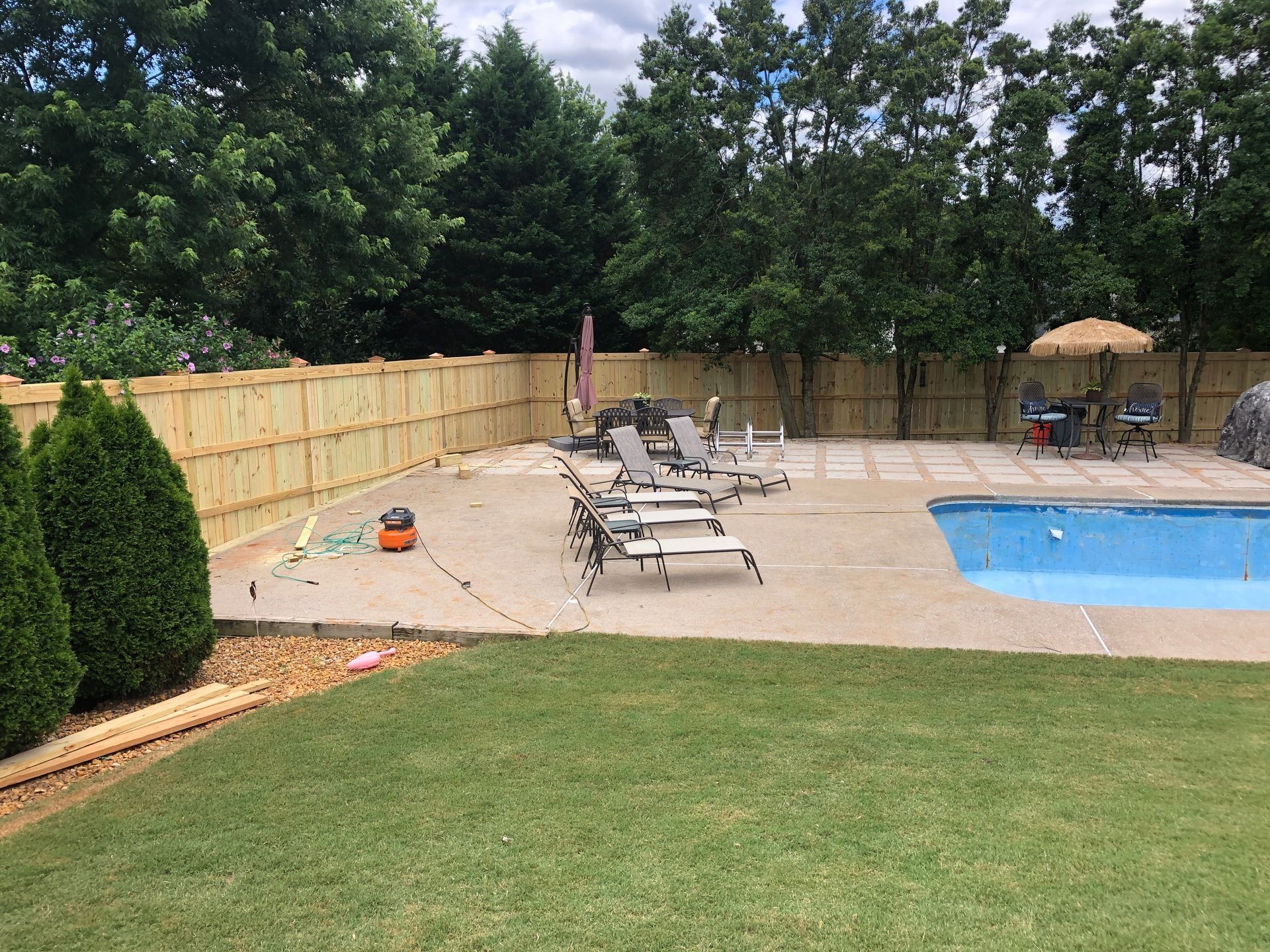 Backyard pool patio with lounge chairs, wooden fence, trees, and green lawn