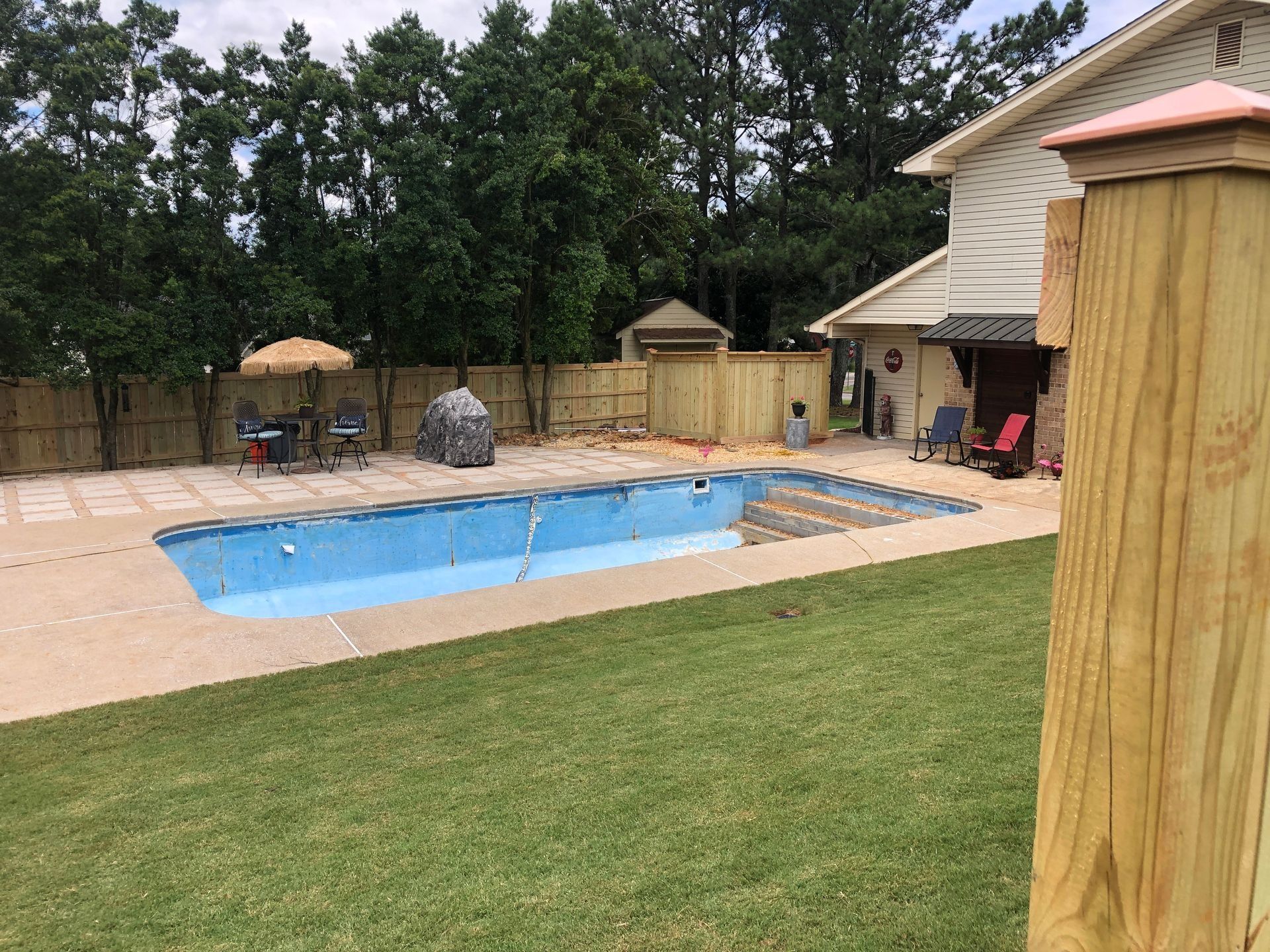 Backyard with an in-ground pool, tan patio, lounge chairs, and a wooden fence under trees