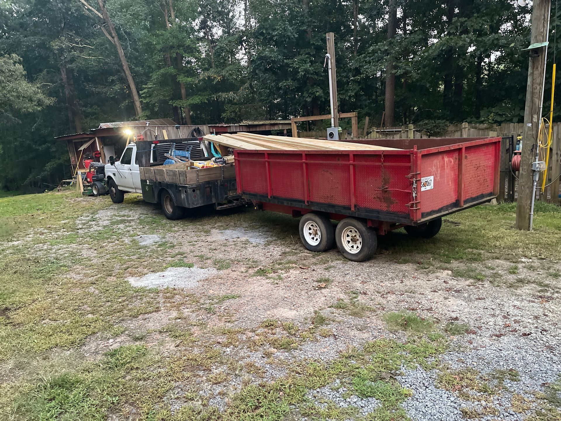 Red dump trailer and a flatbed truck parked on a gravel lot beside trees