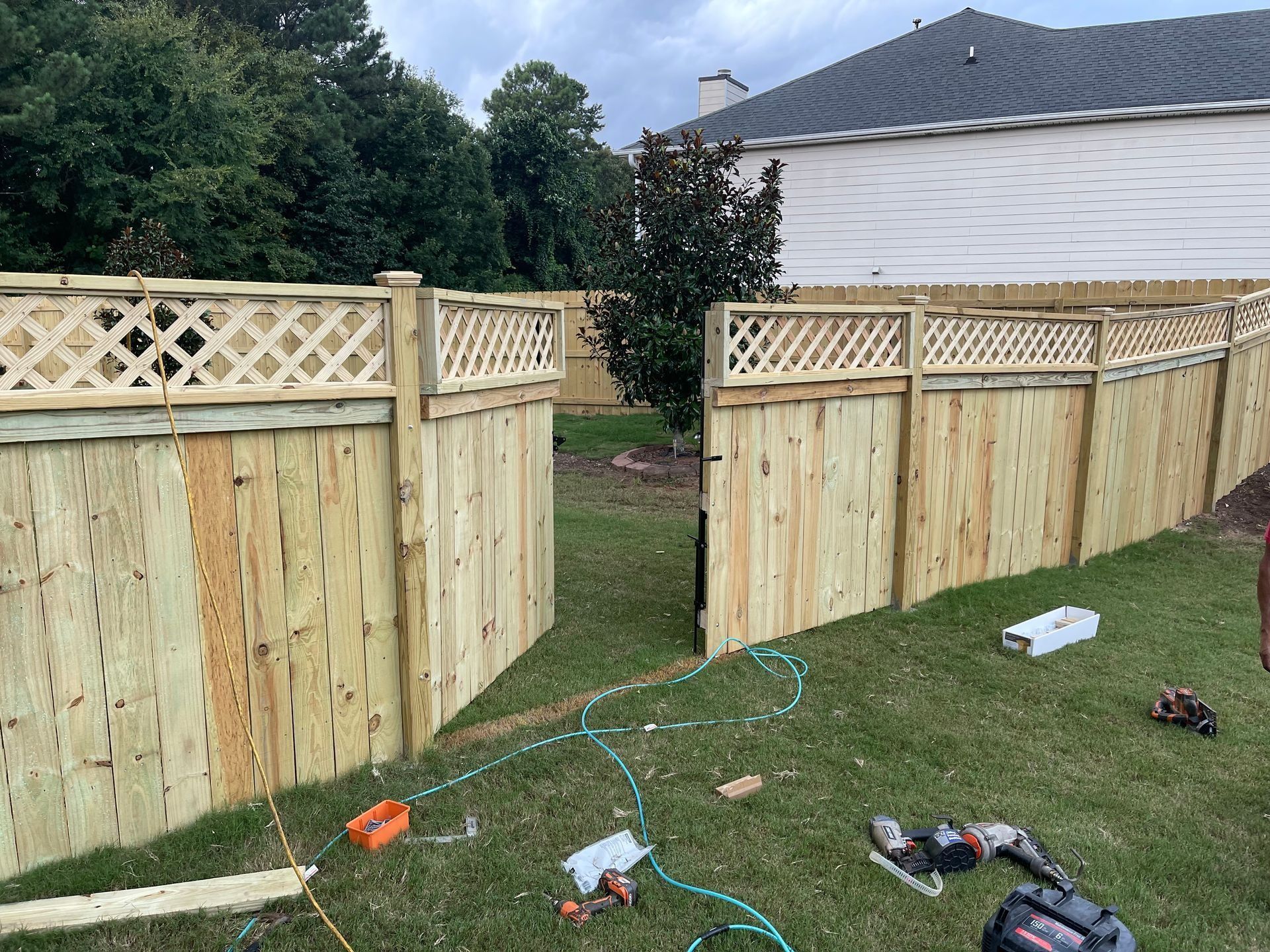 Two wooden privacy fences meeting at a corner in a backyard, with grass and scattered tools in front.