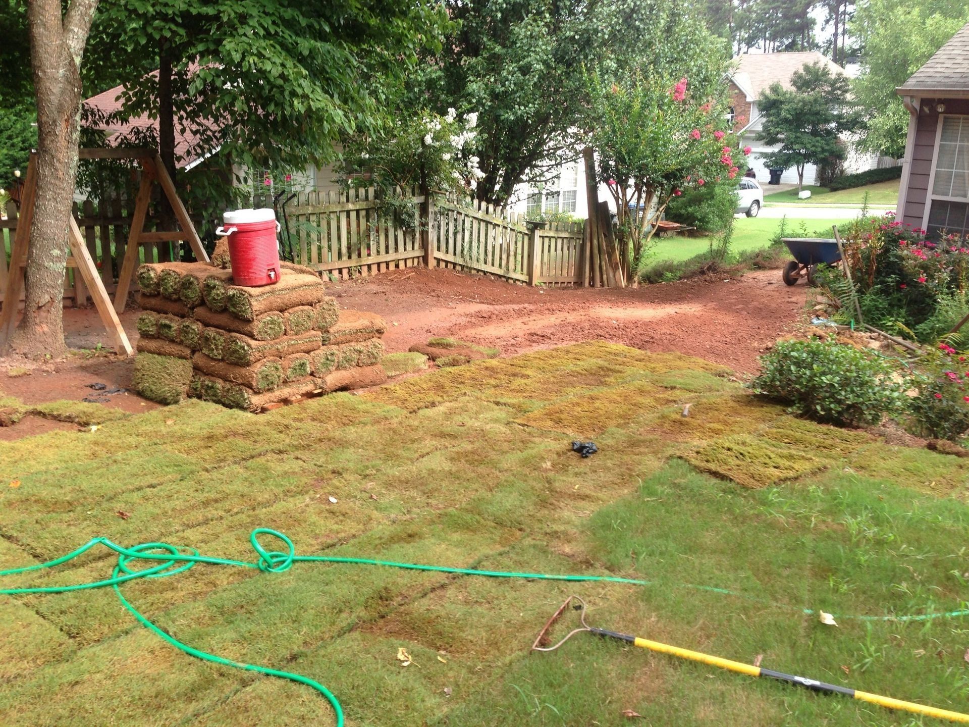 Backyard with a muddy patch, green hose, stacked bricks, and a red bucket near a white fence