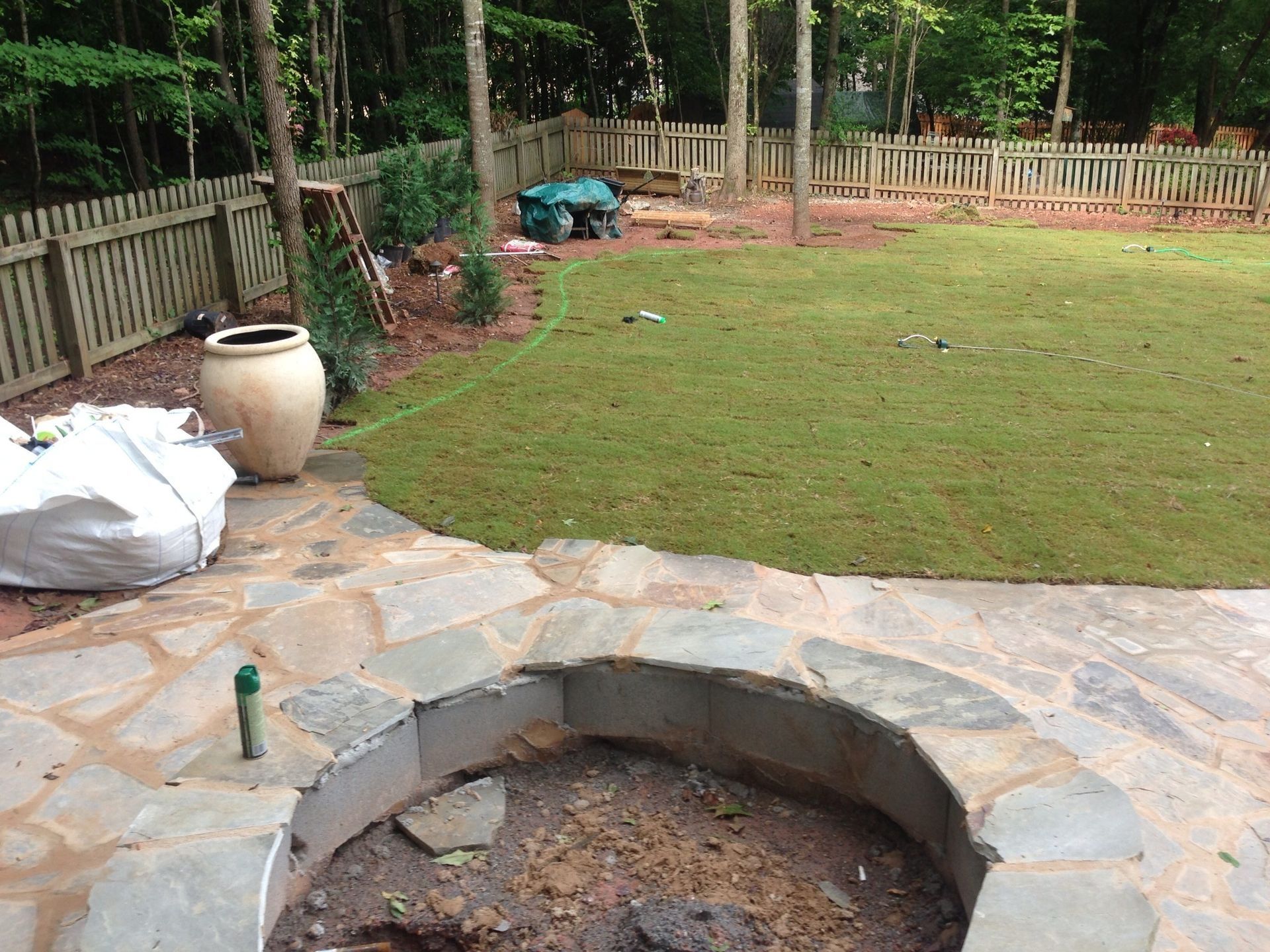 Backyard stone patio with a circular fire pit, grassy lawn, and wooden fence surrounded by trees