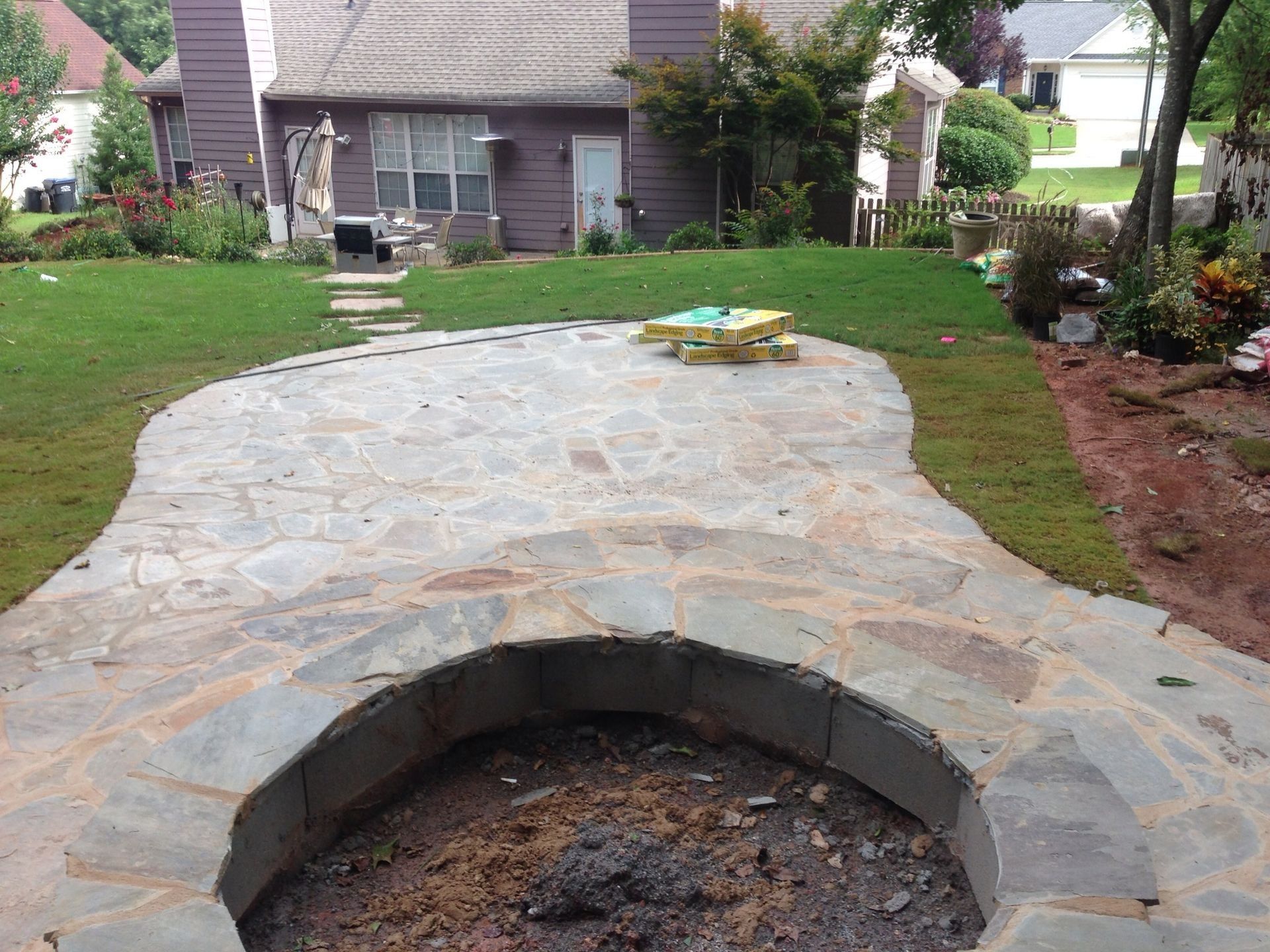 Stone patio with a circular fire pit in a backyard yard, leading to a house and garden beds