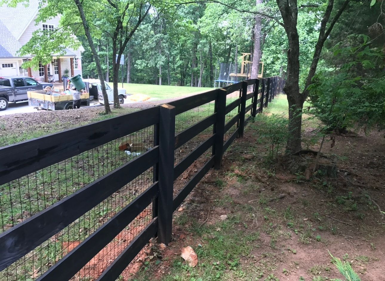 Black wooden fence along a grassy, tree-lined path beside a house and driveway
