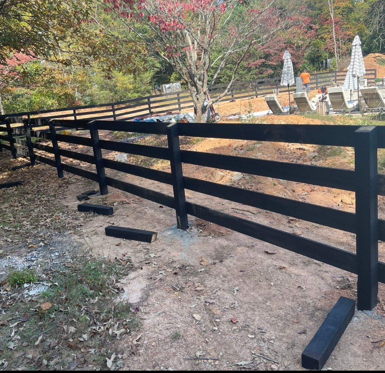 Black wooden fence along a dirt path in a wooded yard, with trees and patio umbrellas in the background