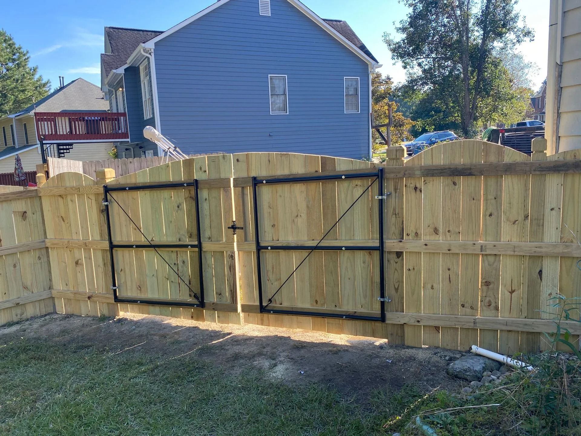 Wooden backyard fence with double gates in front of a blue house, viewed from a grassy yard.