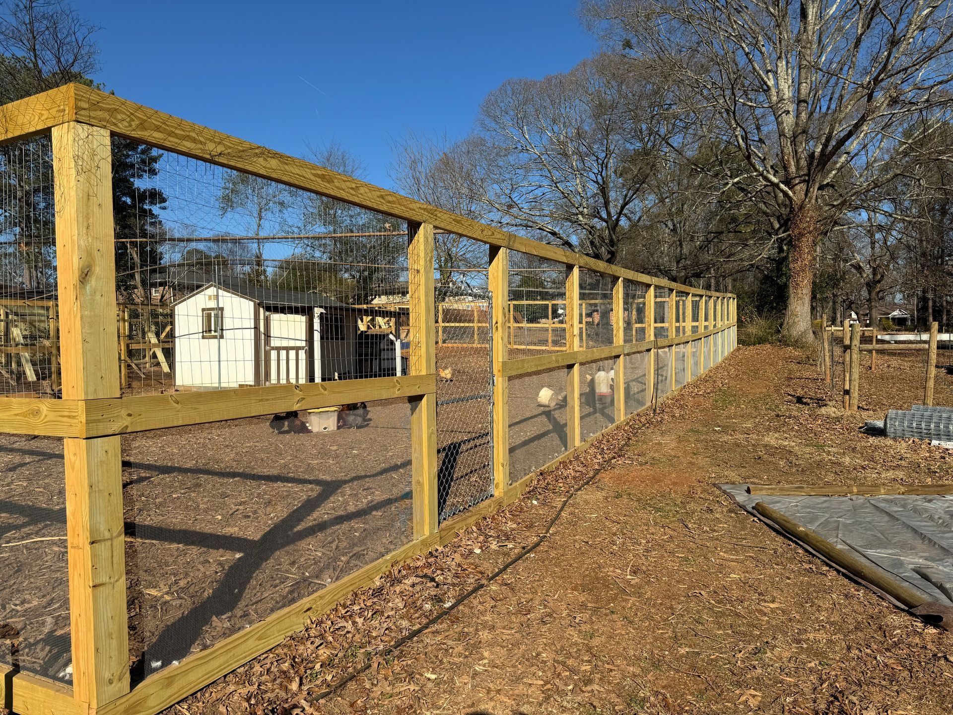 Wooden fence under construction along a dirt path beside a white shed and leafless trees