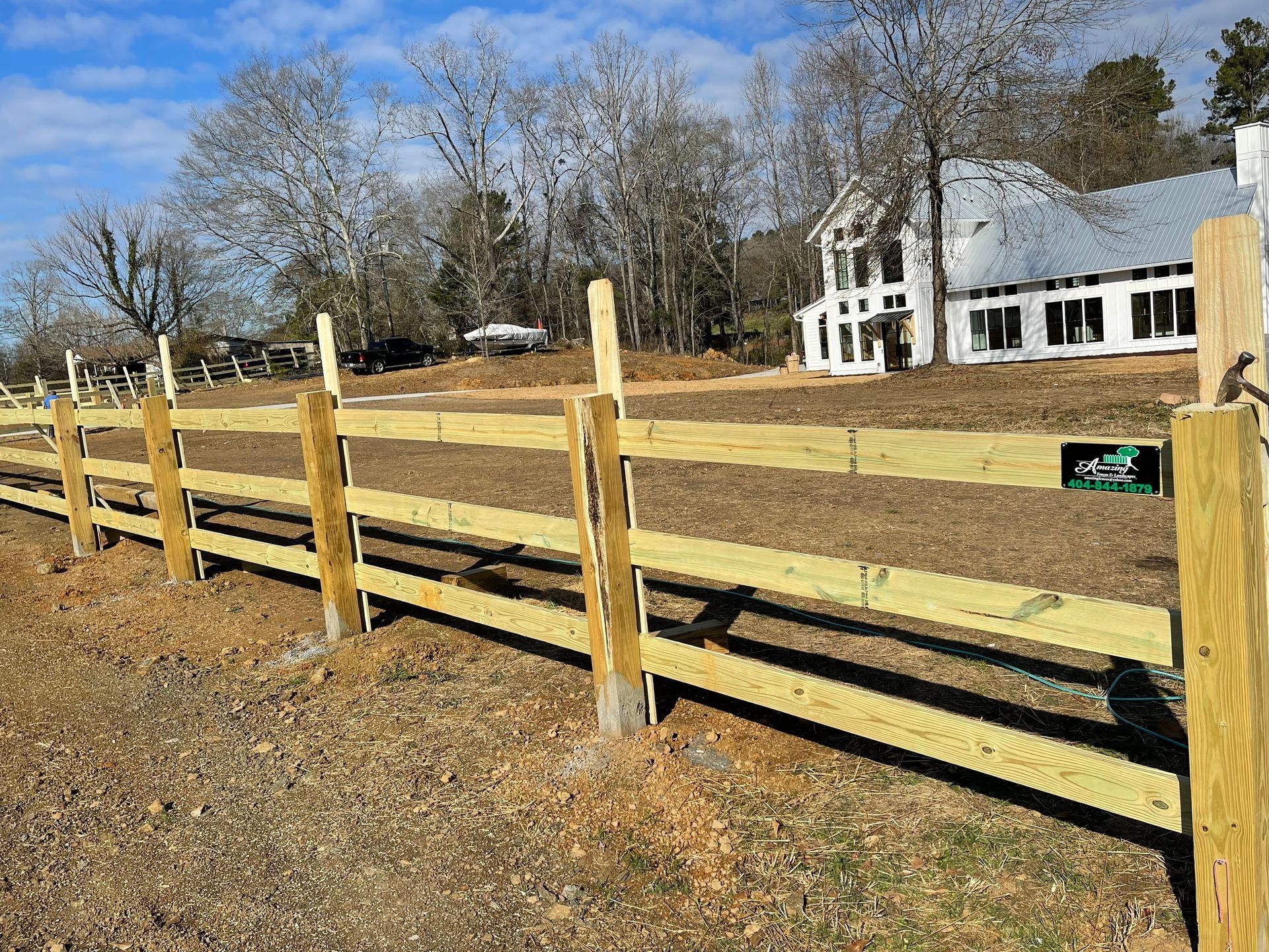 Wooden split-rail fence beside a grassy field and white farmhouse under a clear blue sky