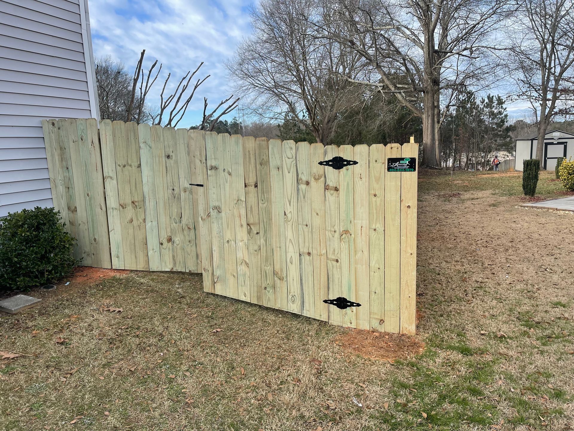 Wooden backyard gate beside a house, with black hinges and a dirt path leading past it