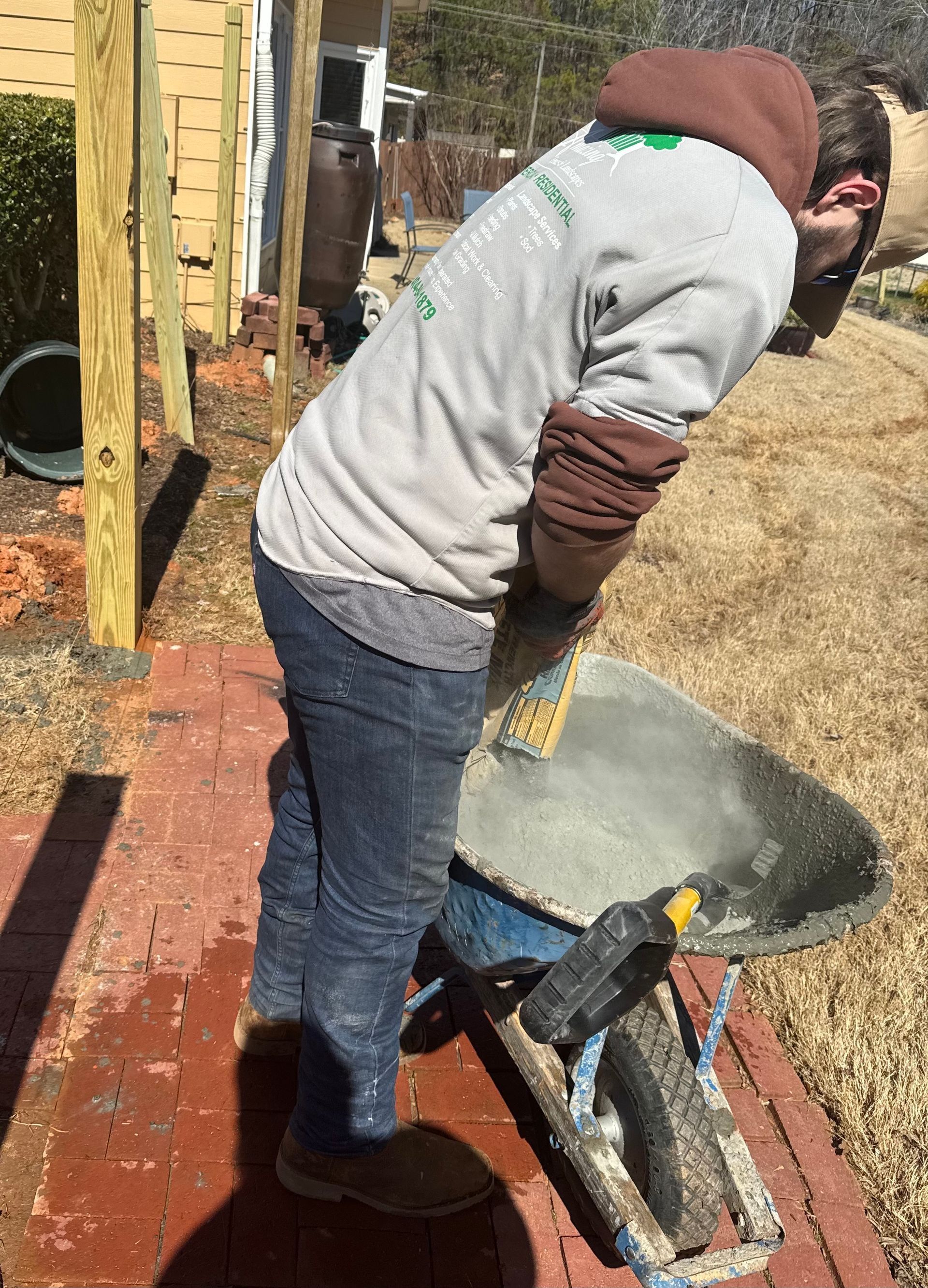 Person bending over a wheelbarrow outdoors beside a brick path and wooden fence.