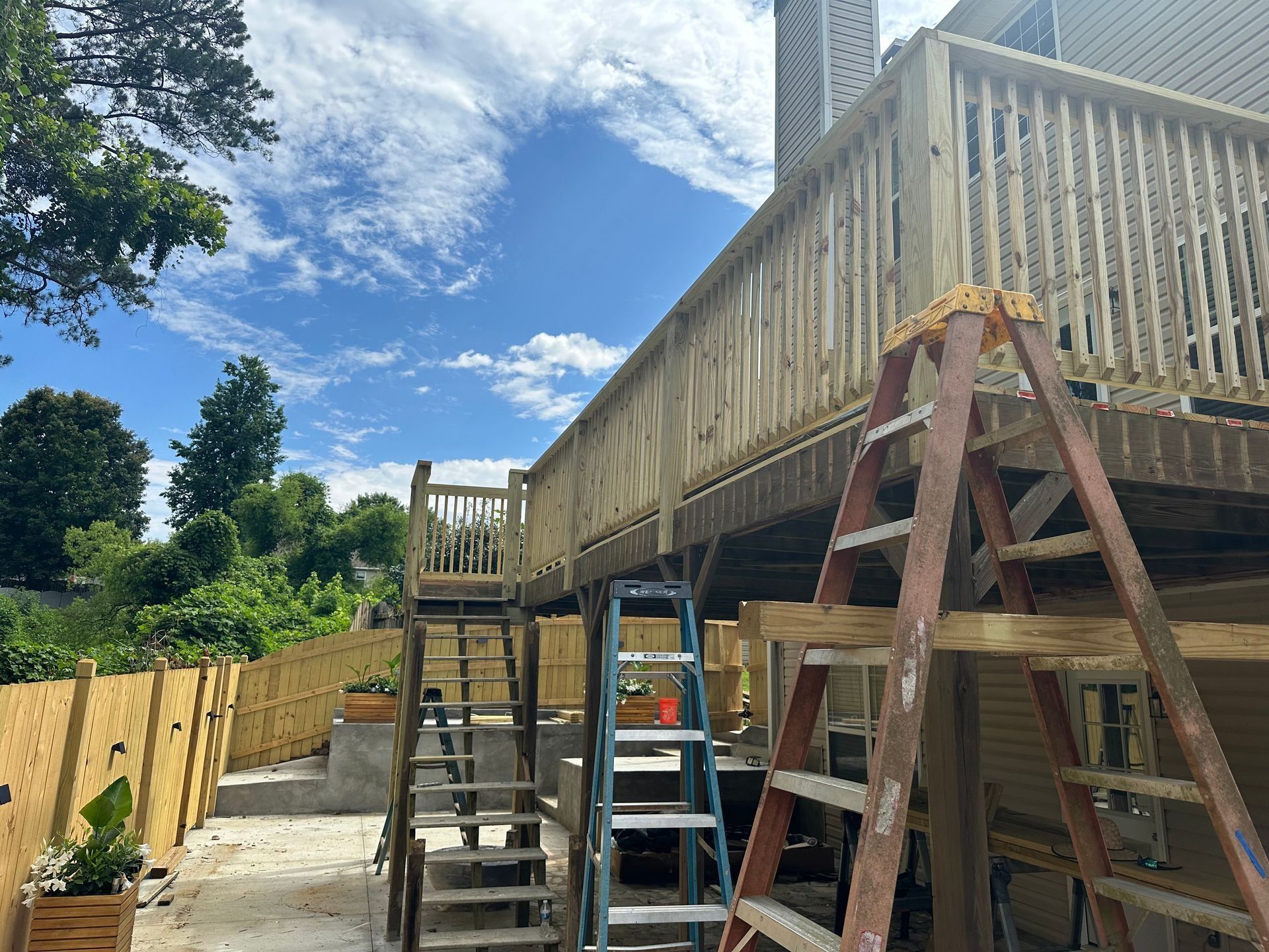 Wooden deck under construction with ladders, railings, and a cloudy blue sky above the yard