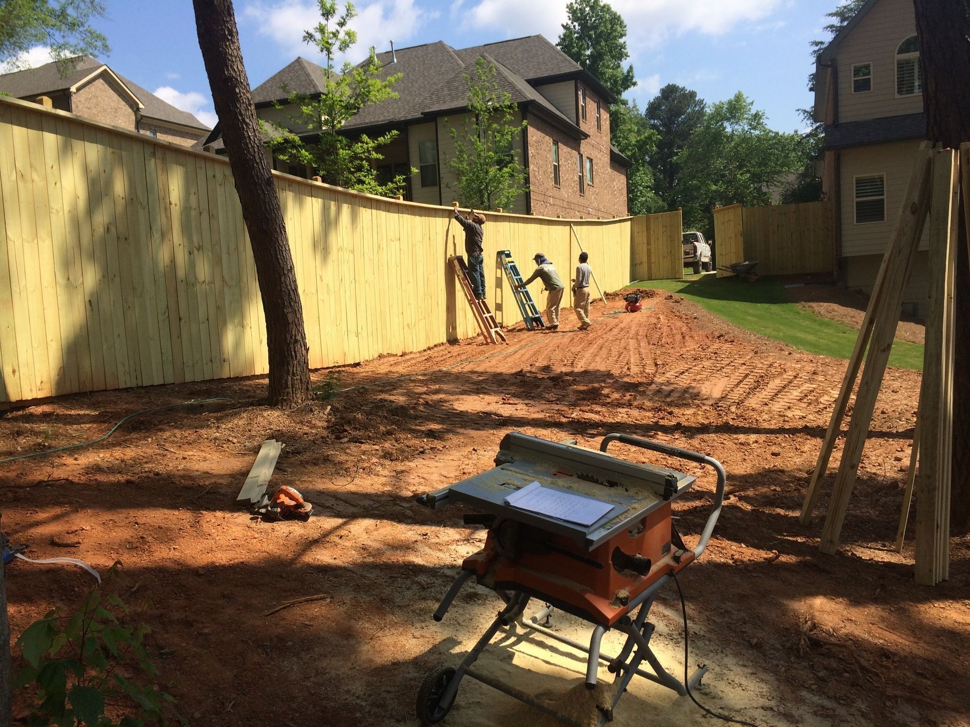 Backyard under construction with dirt ground, yellow fence, houses, workers, and a wheelbarrow in foreground