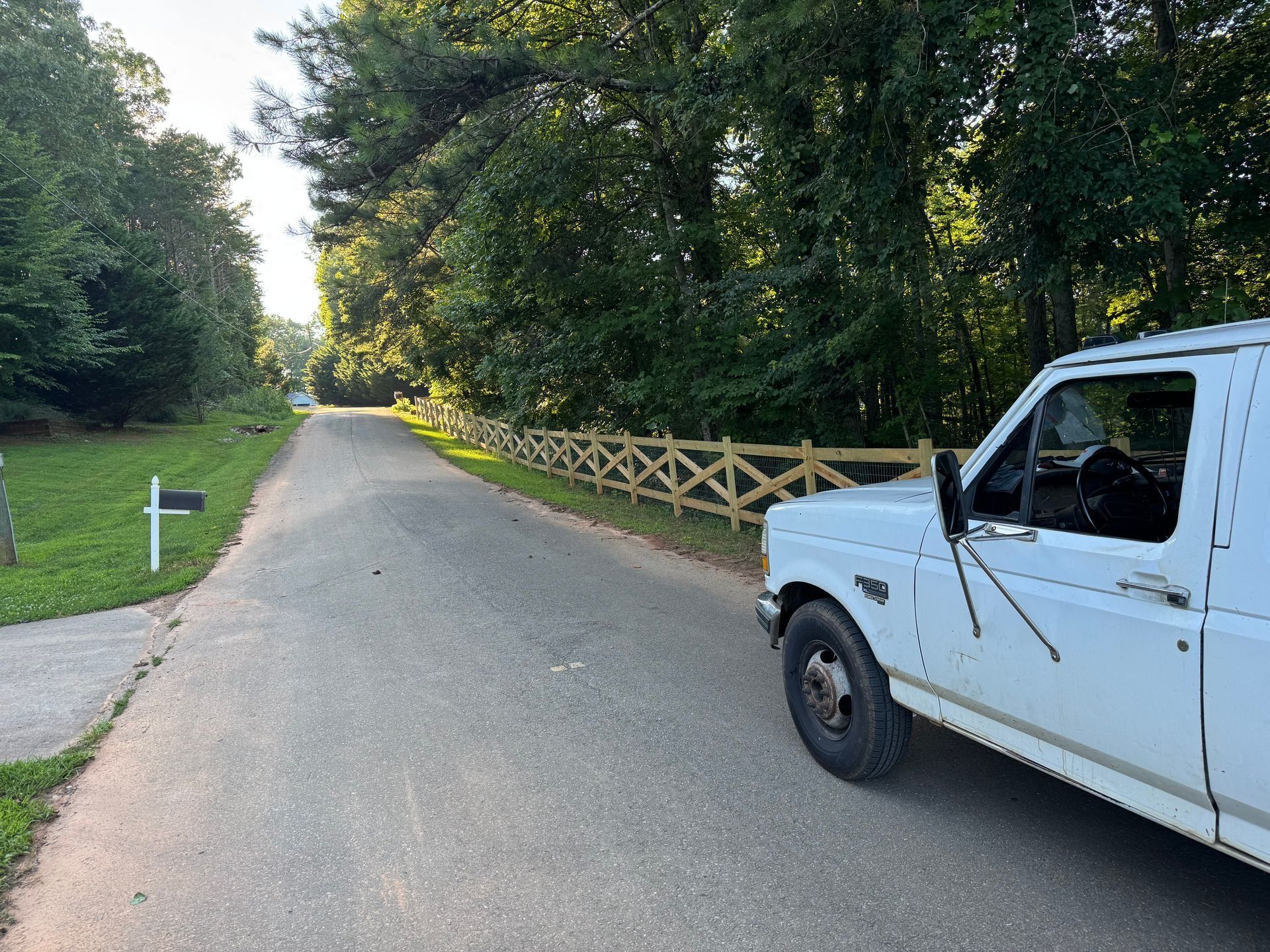 White pickup truck on a quiet tree-lined road beside a wooden fence.