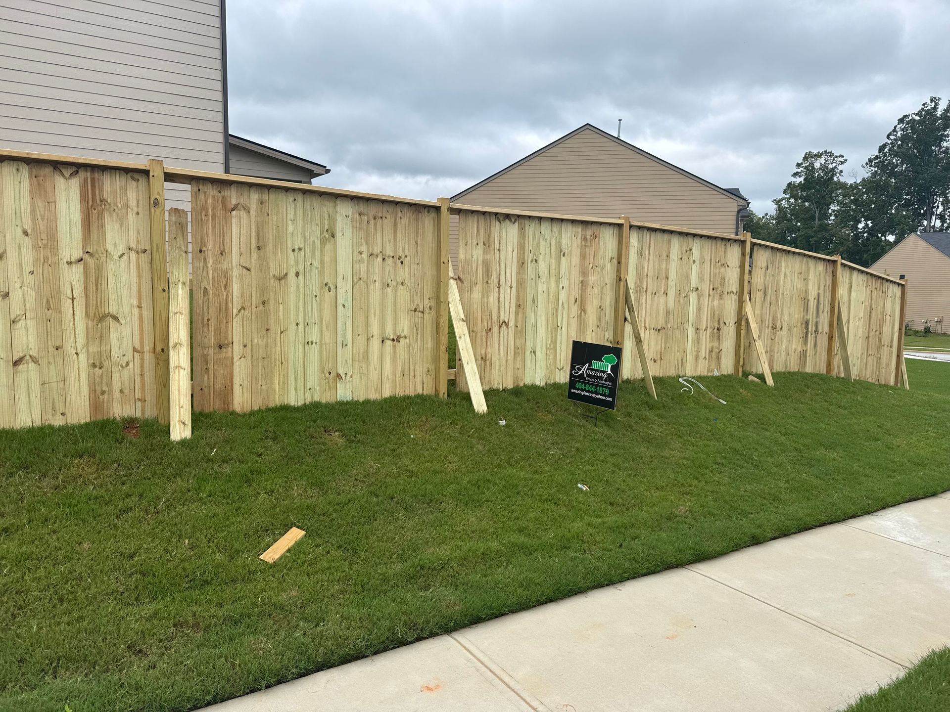Backyard with a long wooden fence, green lawn, and sidewalk under cloudy skies