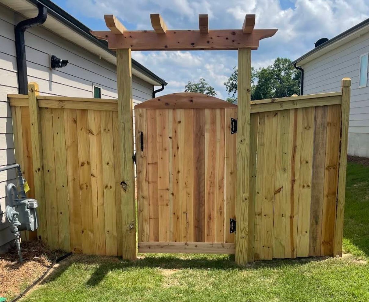 Wooden backyard privacy fence with a central gate and pergola, surrounded by grass and neighboring houses.