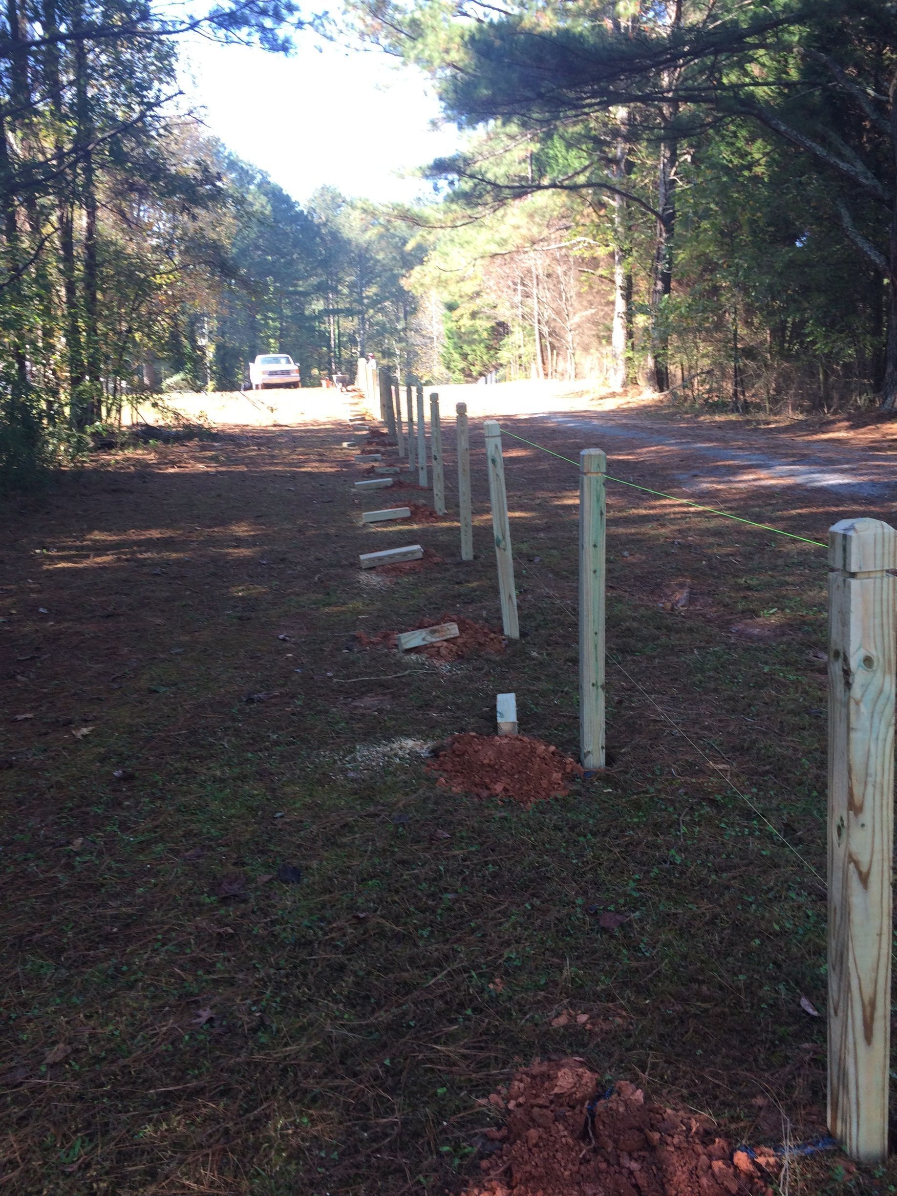 Wooded path lined with white fence posts and red dirt patches, sunlit at the far end