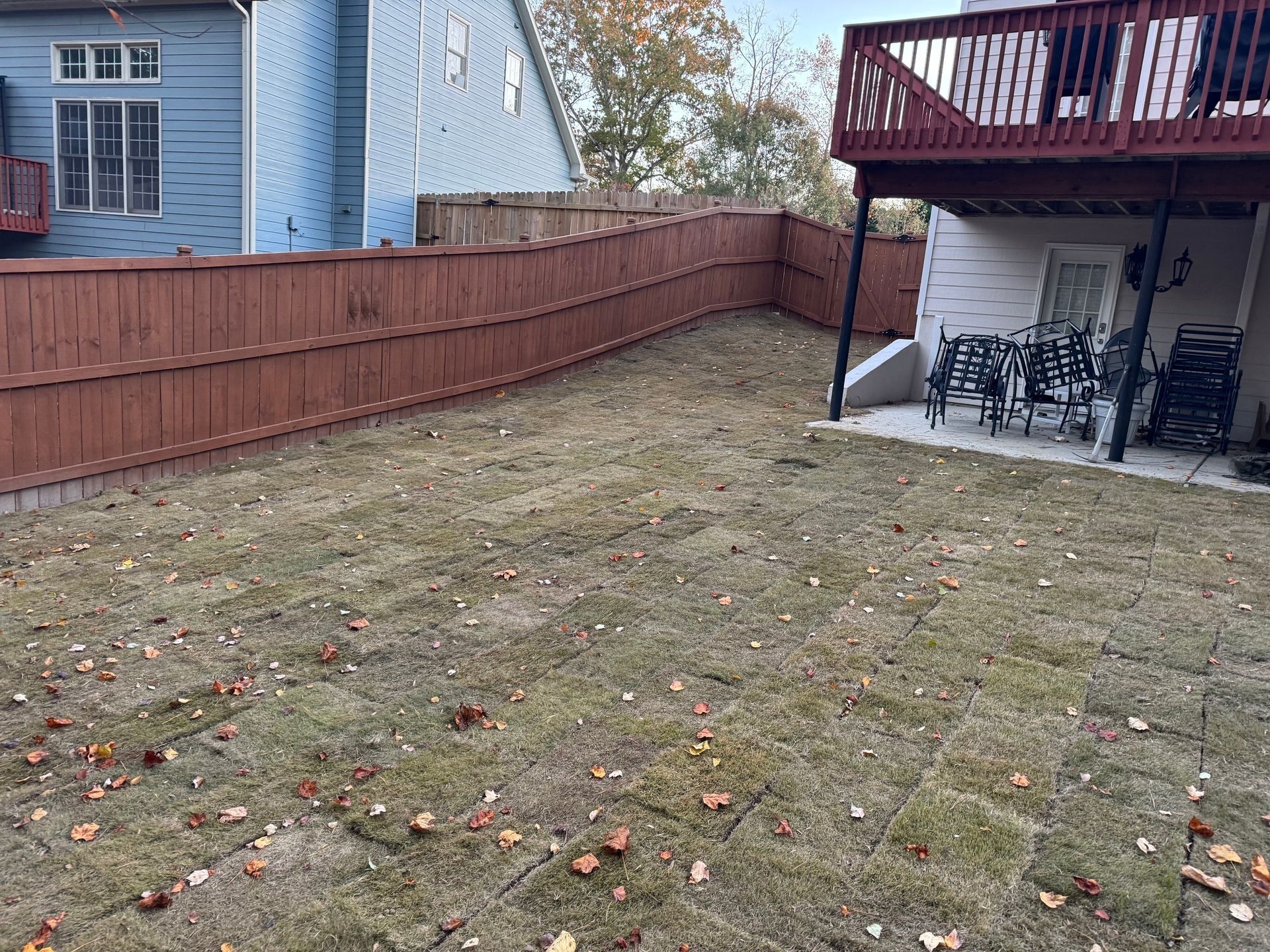 Backyard with patchy grass, red wooden fence, and covered patio with chairs and grill