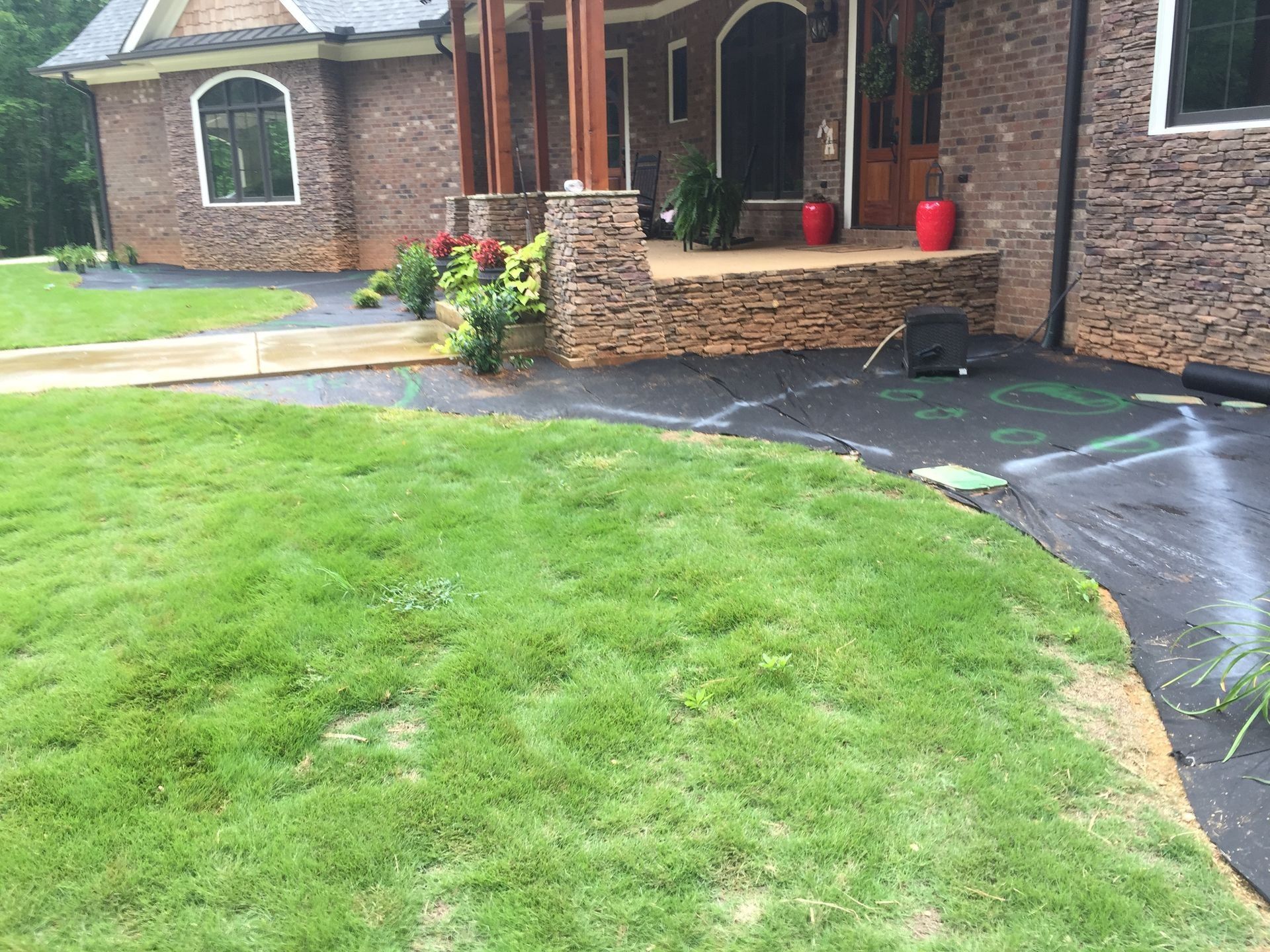 Brick house with covered porch, stone planter, and wet curved driveway beside a green lawn.