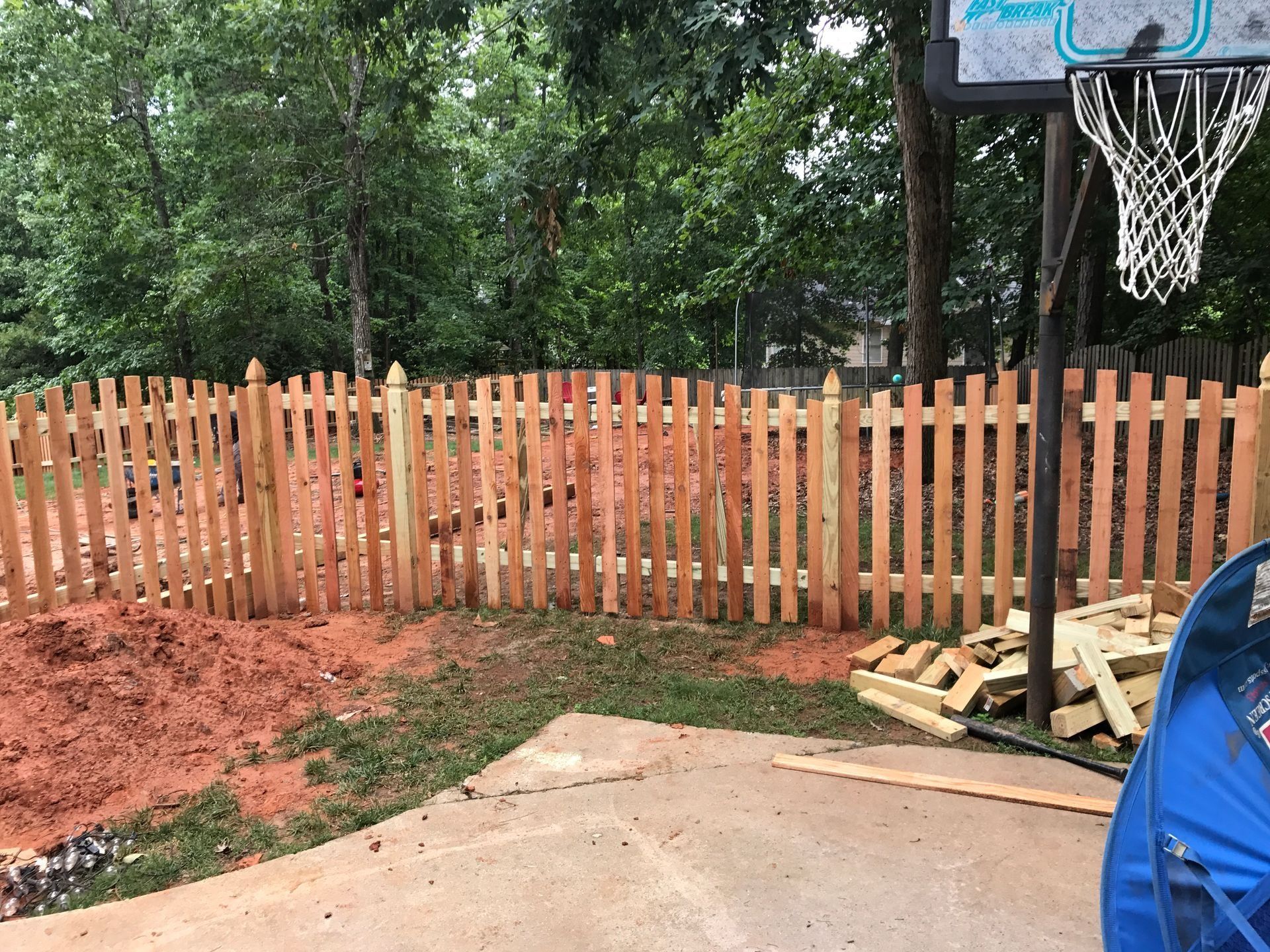 Wooden fence along a muddy yard beside a basketball hoop and blue tarp in a wooded area