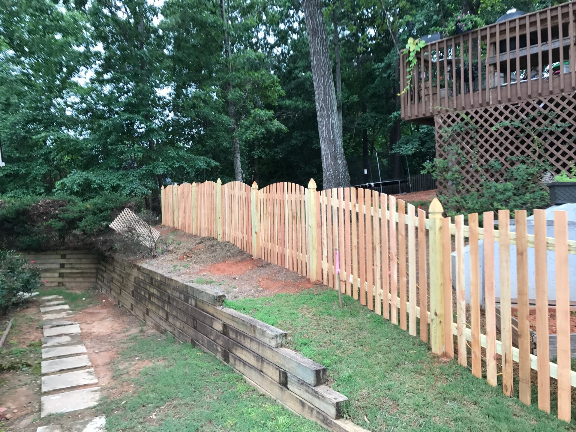 Backyard with a wooden fence, stone path, and brick retaining wall beside a grassy slope and trees