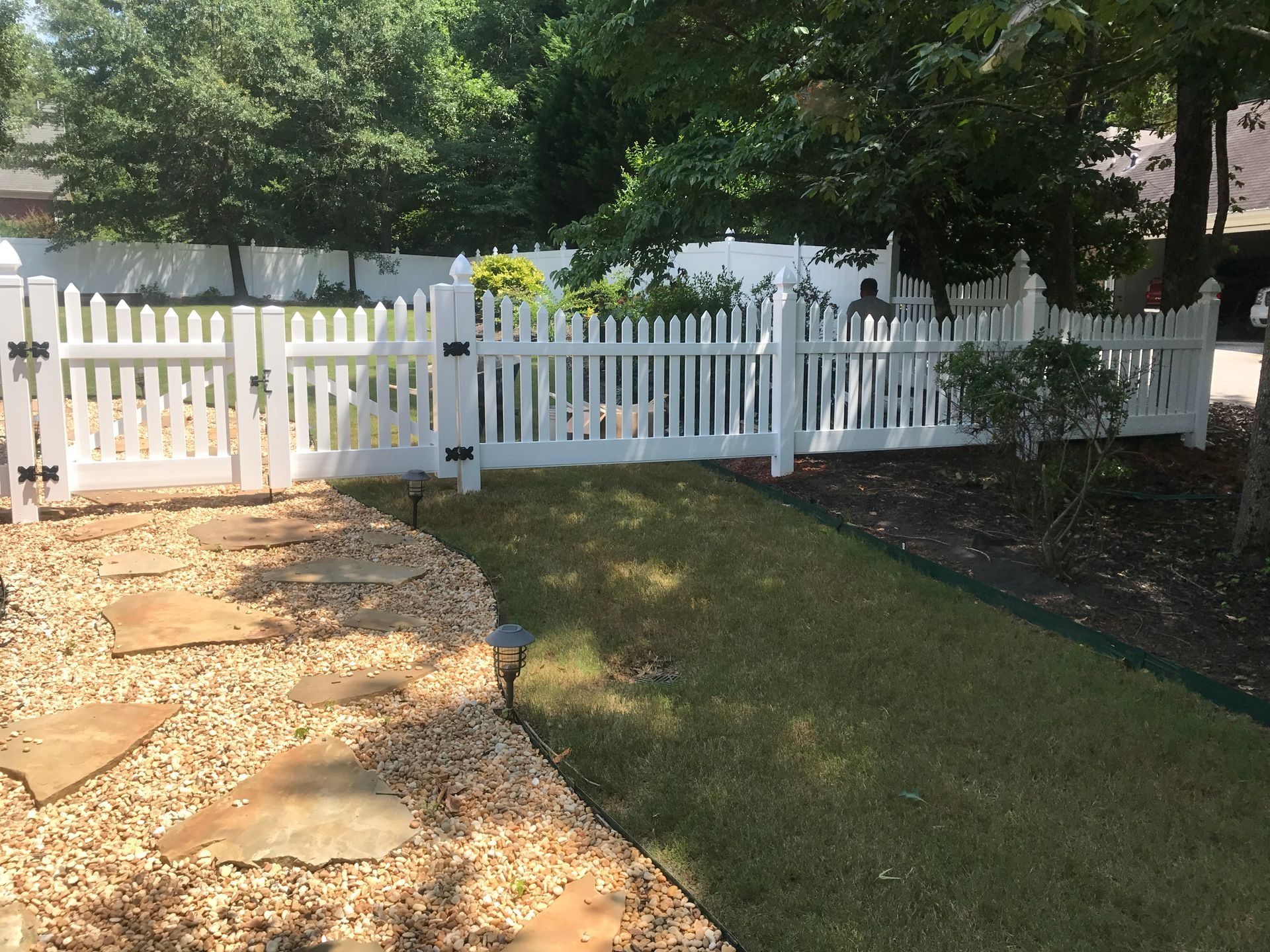 White picket fence separating a grassy yard from a stone path and mulch bed under trees