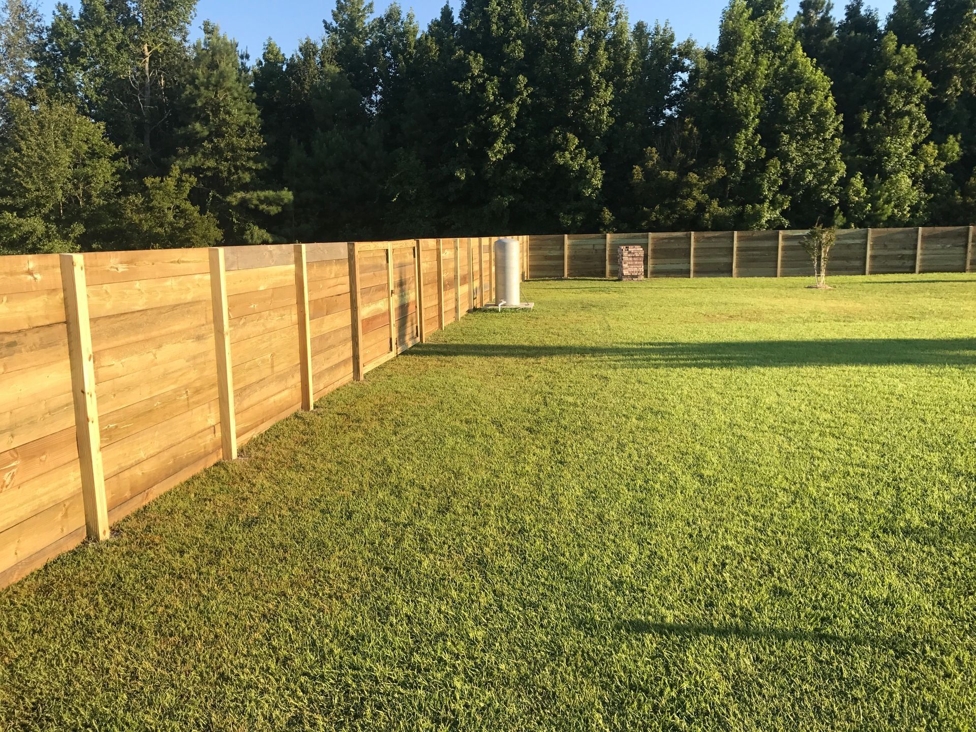 Large grassy backyard with a long wooden fence and trees in the distance.