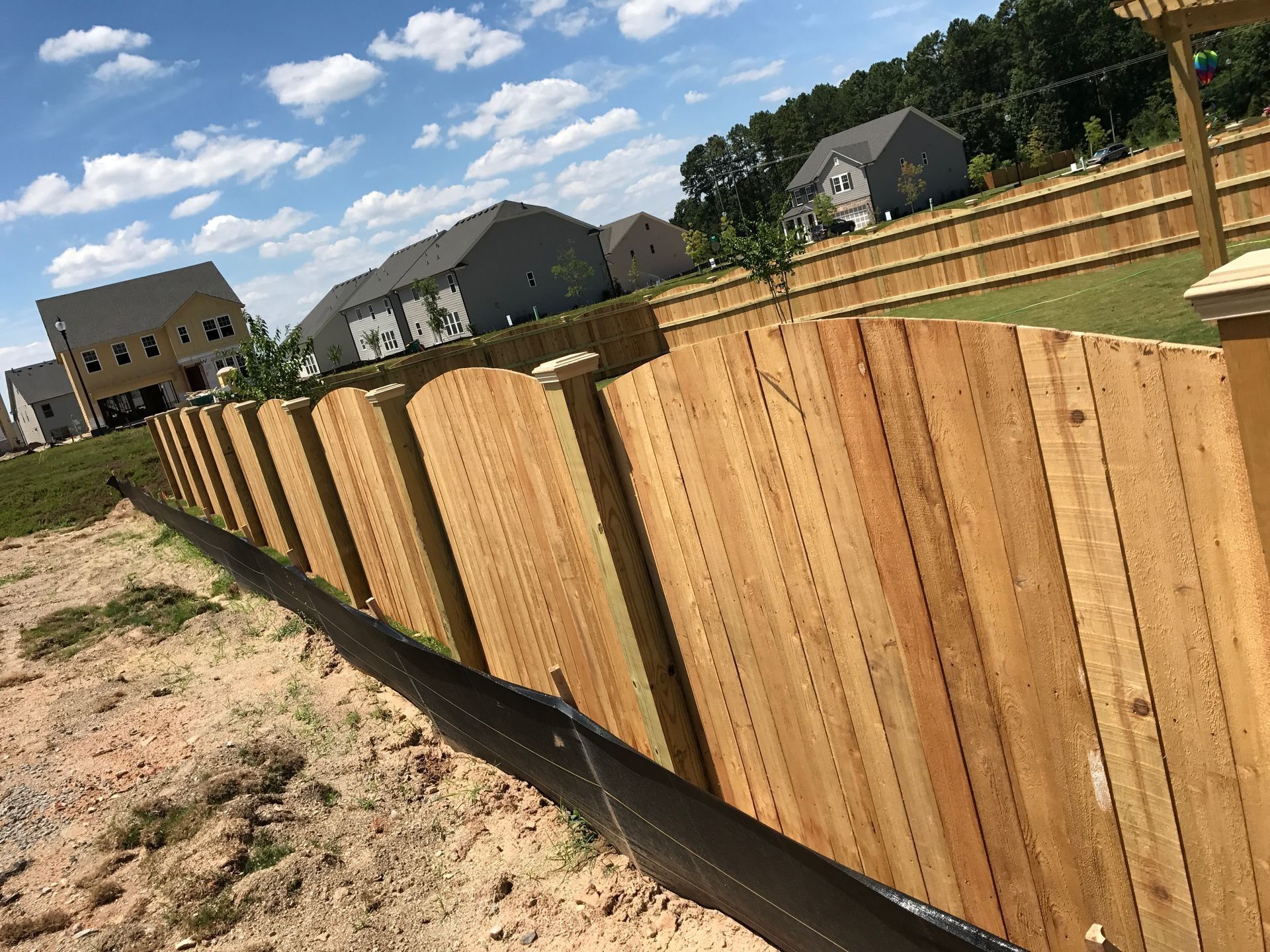 New wooden privacy fence along a sandy backyard under a blue sky with houses in the background