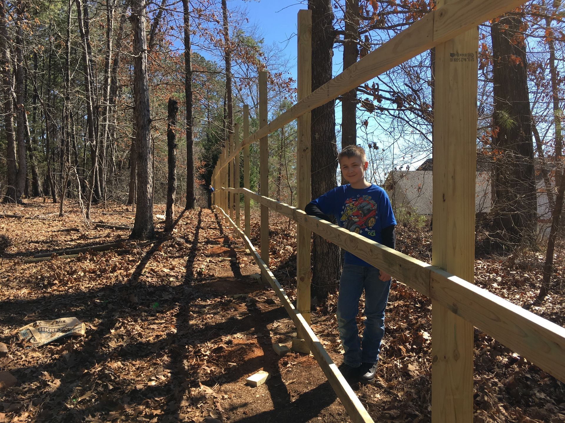 Person in a blue shirt walking beside a wooden fence in a sunlit forest.