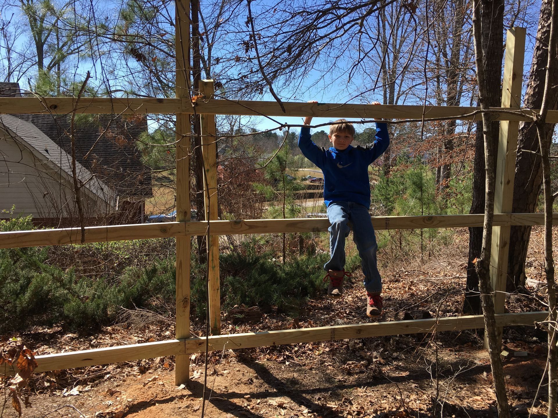 Person in a blue jacket hanging from a wooden fence outdoors in a wooded yard.
