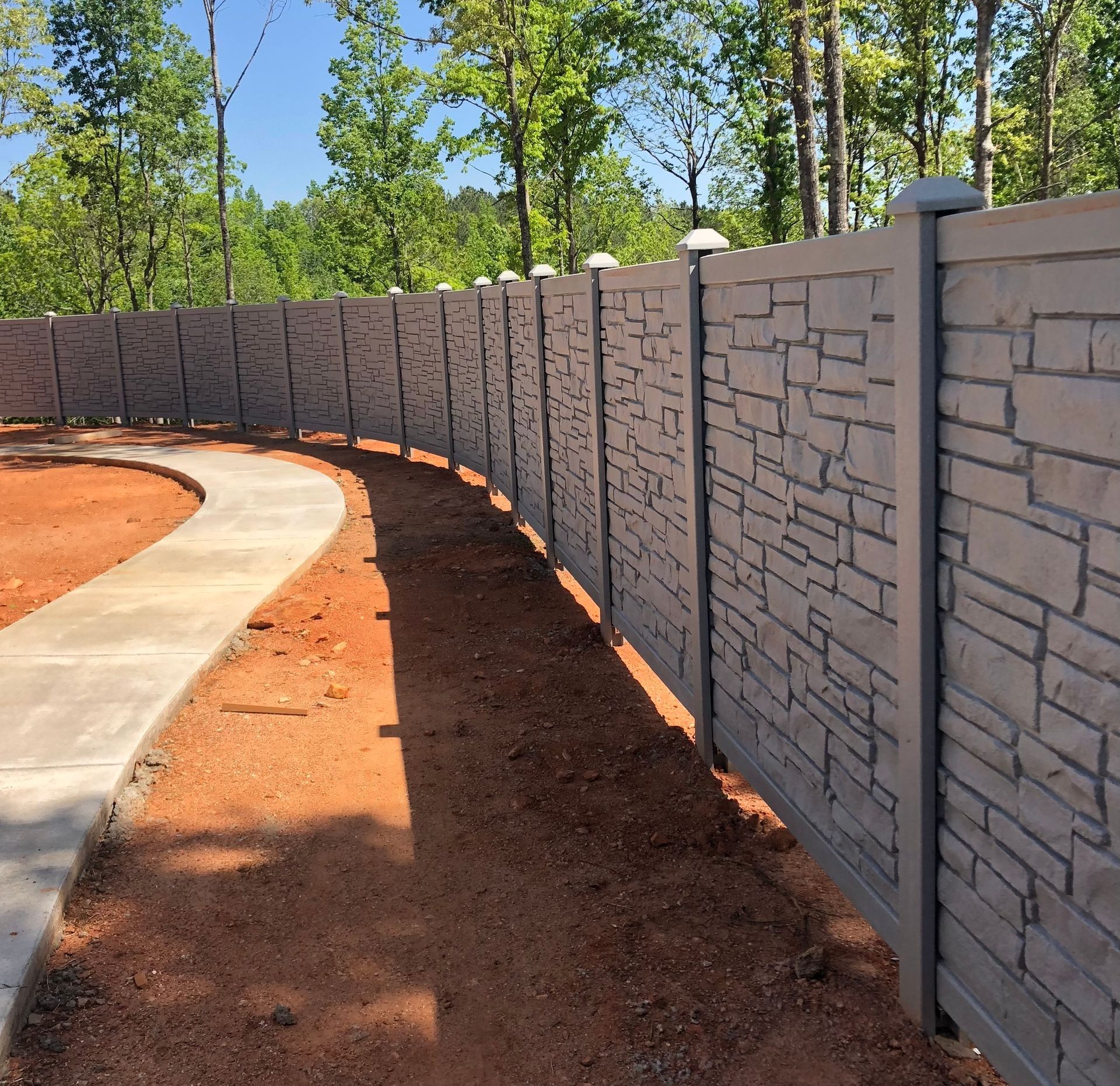 Curved concrete path beside a gray stone privacy wall with trees and mulch along the edge