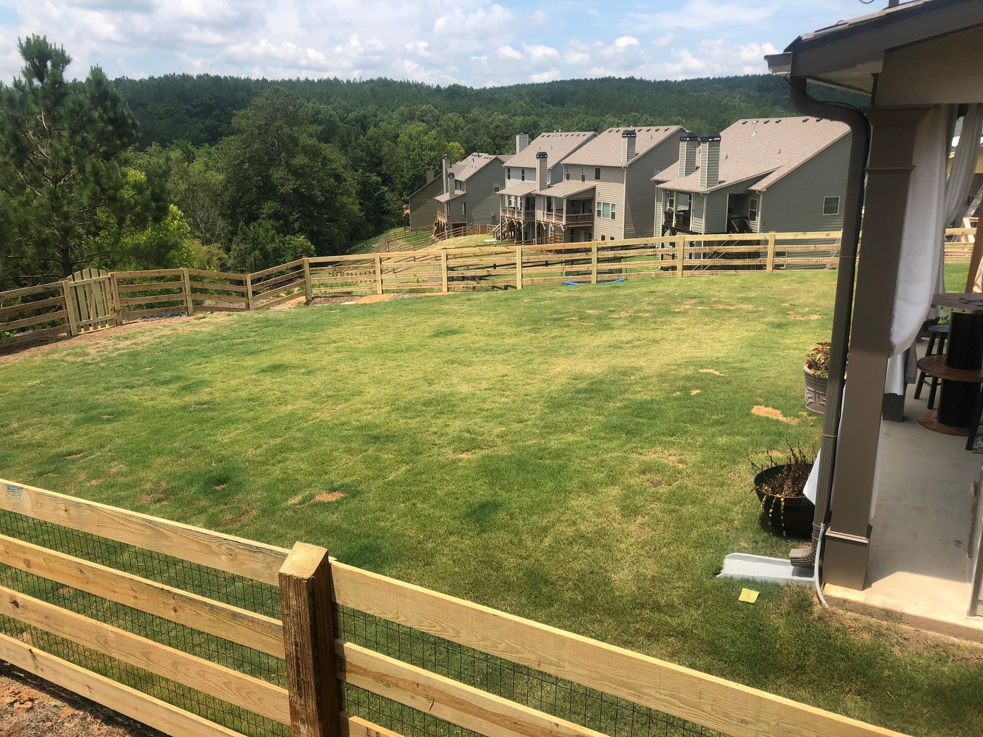 Backyard lawn with wooden fence, patio, and apartment buildings on a hillside under a cloudy sky