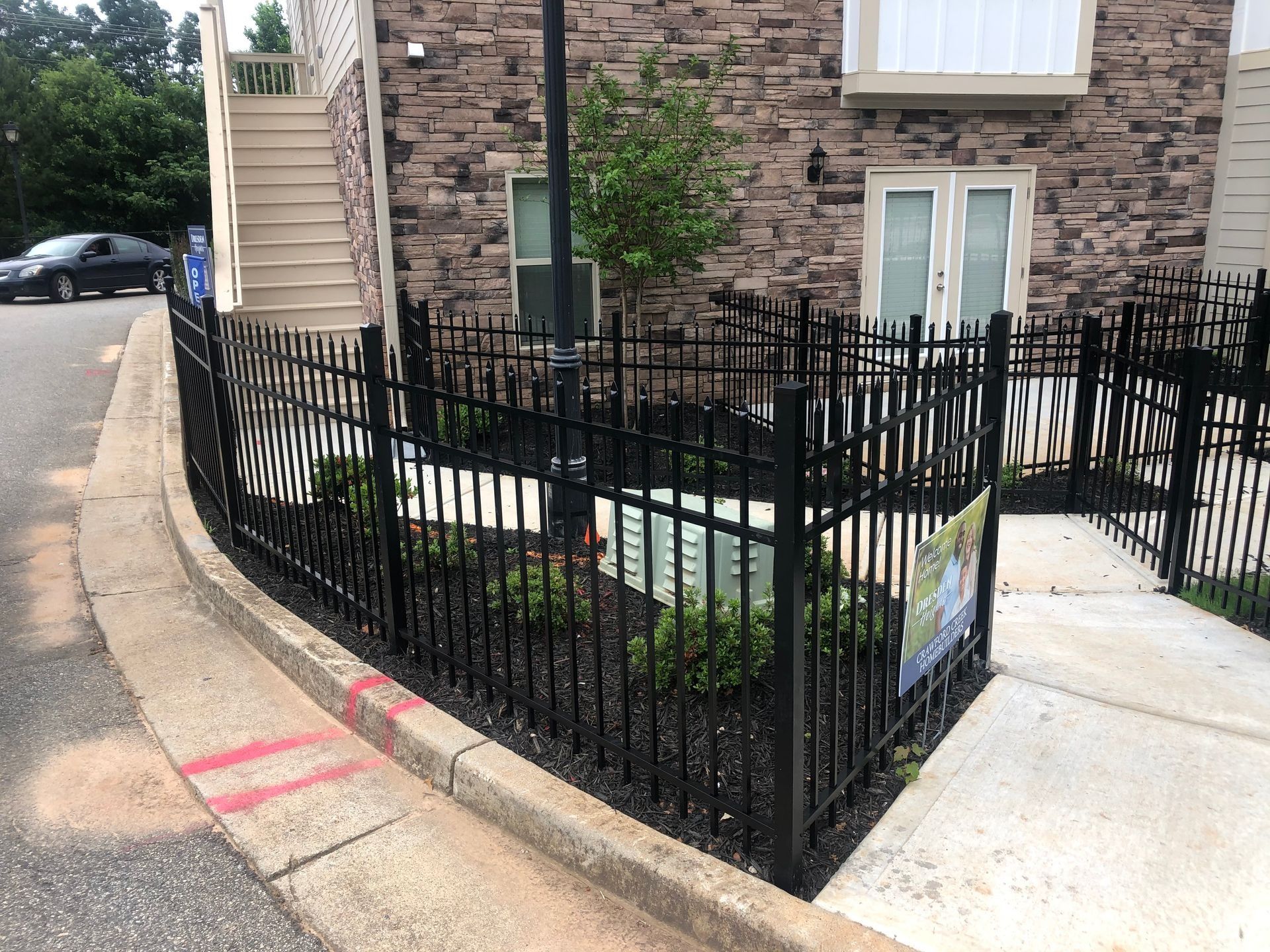 Black metal fence around a small landscaped corner beside a brick building and sidewalk.