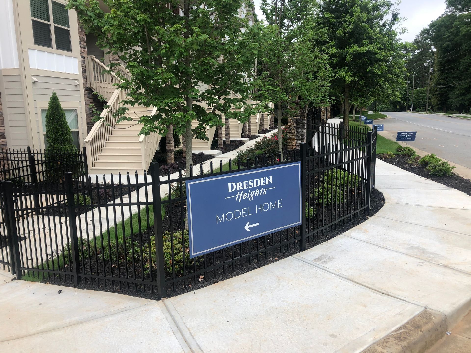 Blue sign on black iron fence outside apartment building beside a sidewalk and street.
