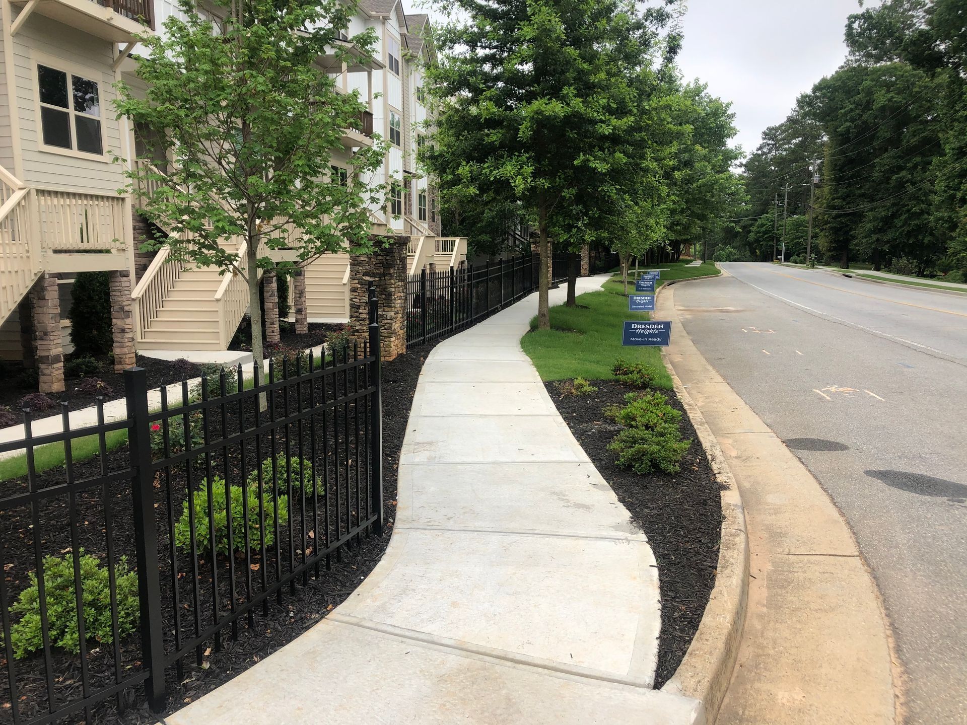 Curved sidewalk beside apartment buildings, black iron fence, and empty road lined with trees and grass.