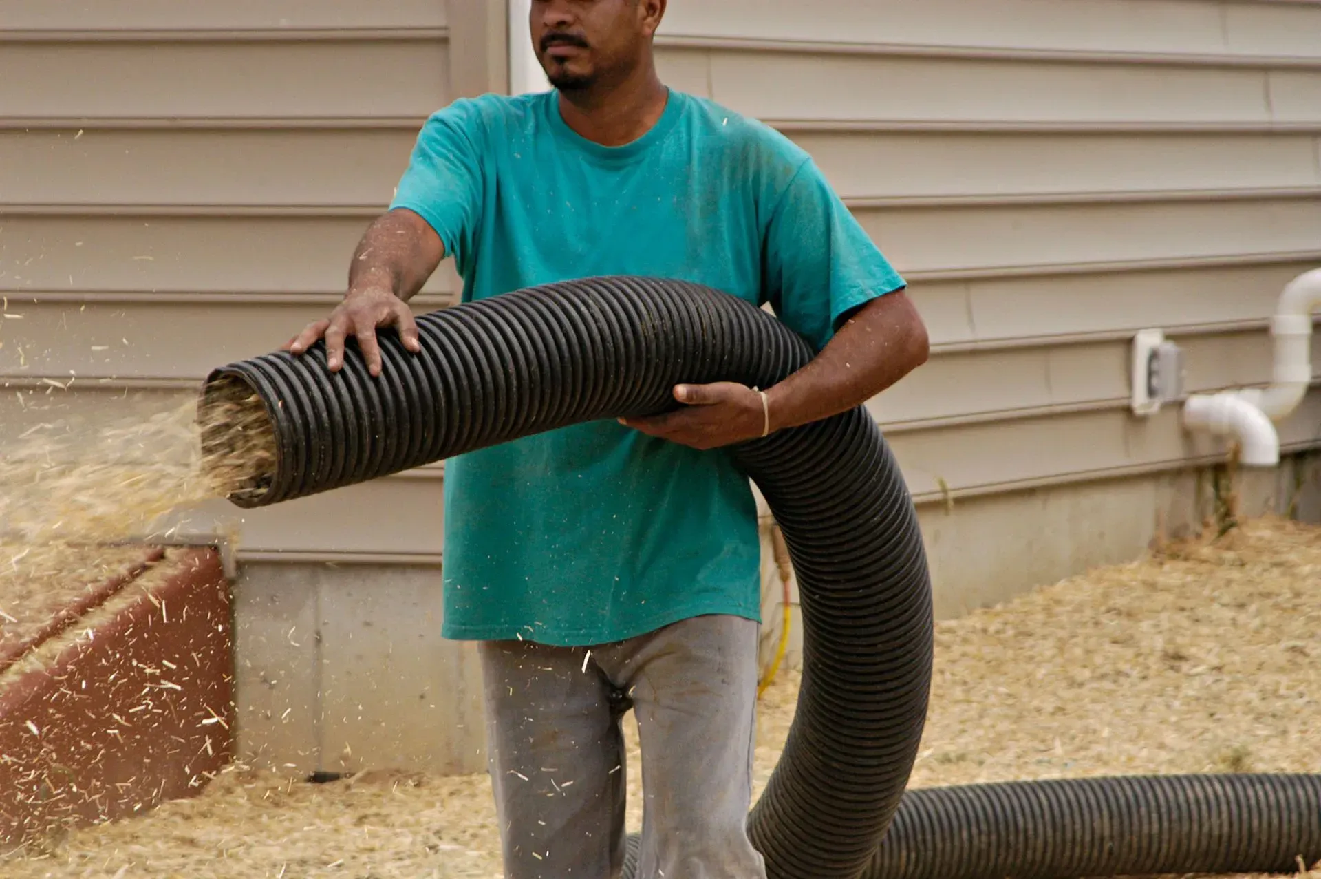 Man holding a large black corrugated hose while spraying water outside a house