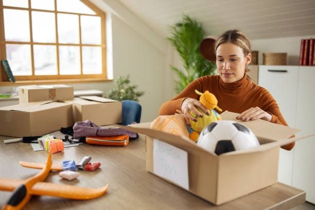 A woman is packing toys into a cardboard box on a table for decluttering before moving.