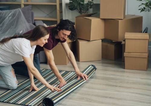 A man and a woman rolling out a carpet in a room full of boxes.