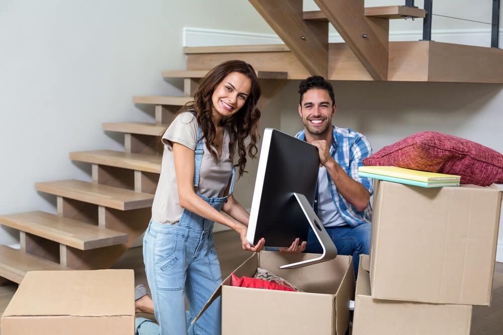 Couple unpacking boxes near a staircase. Woman holds a computer monitor, both smiling. Boxes stacked.