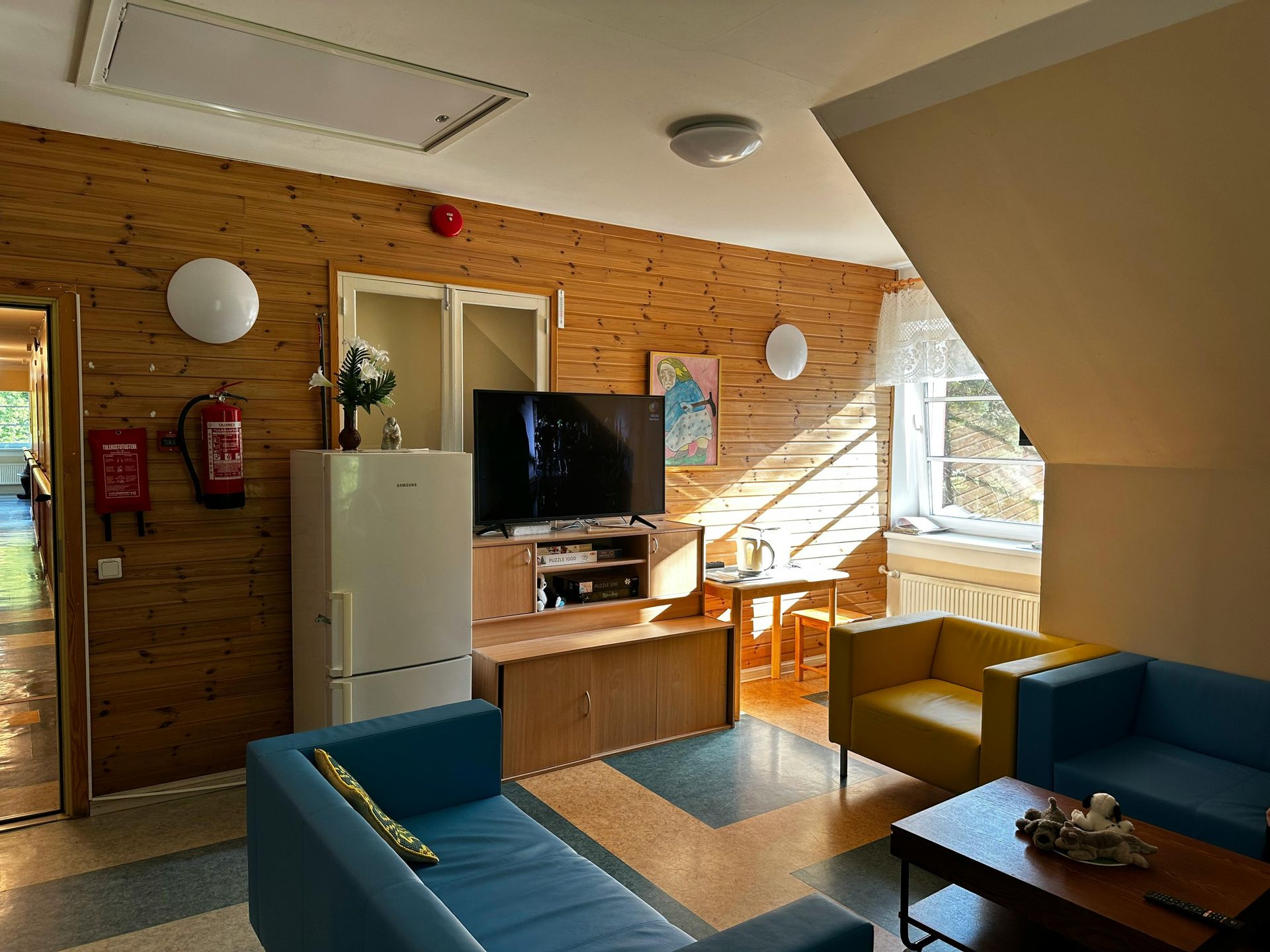 Living room with wood paneling, blue and yellow sofas, TV, and a refrigerator.