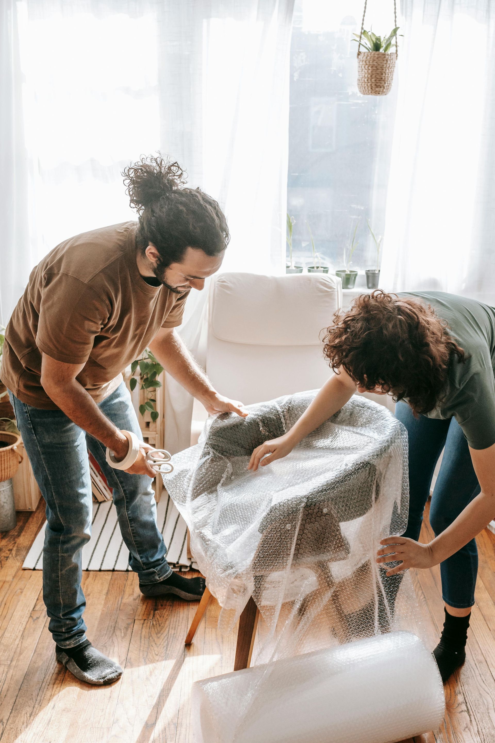 Two people wrapping a chair in bubble wrap inside a sunlit room; smiling.