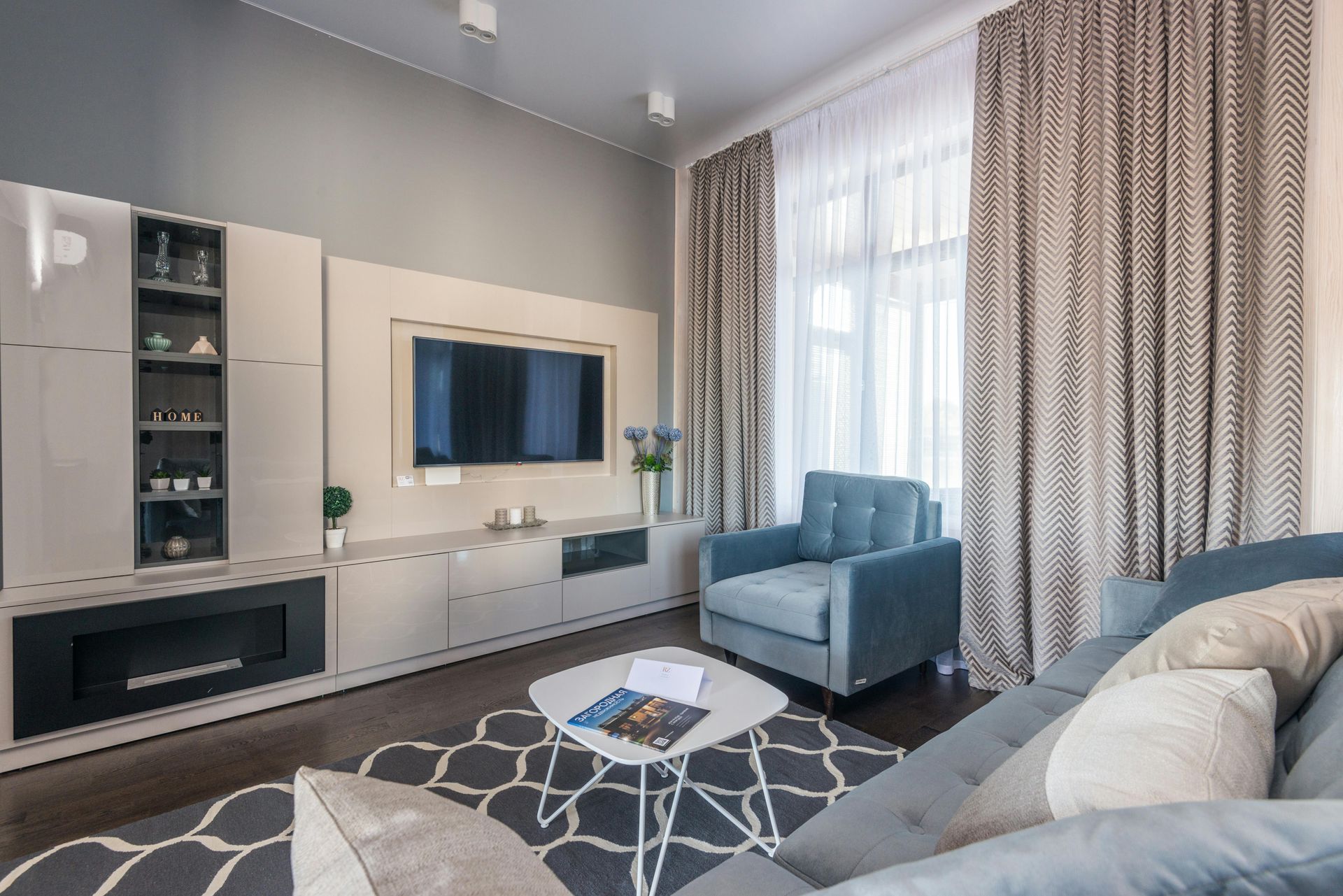 A modern living room with a TV, fireplace console, blue armchair, gray sofa, and geometric rug near floor-length curtains.
