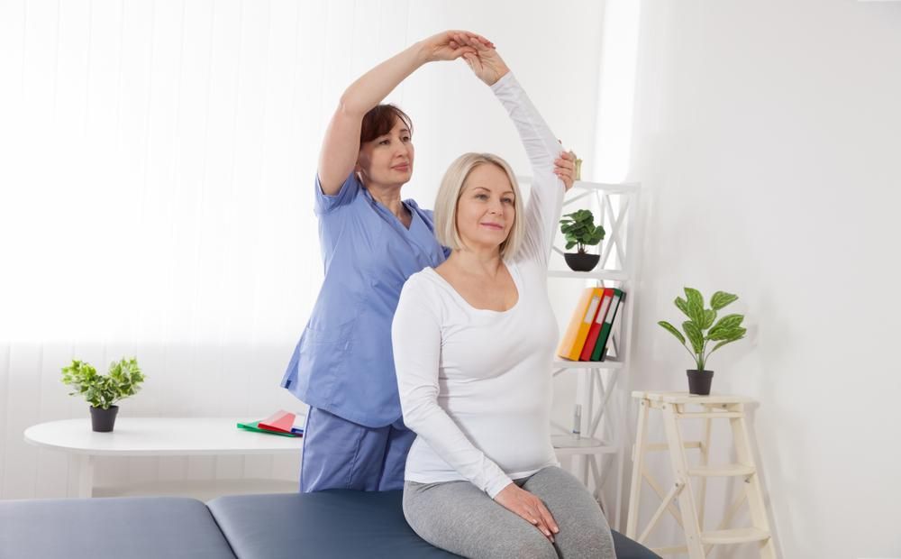 A Woman Is Sitting On A Table While A Nurse Stretches Her Arm — Port Macquarie Physio & Pilates In Port Macquarie, NSW