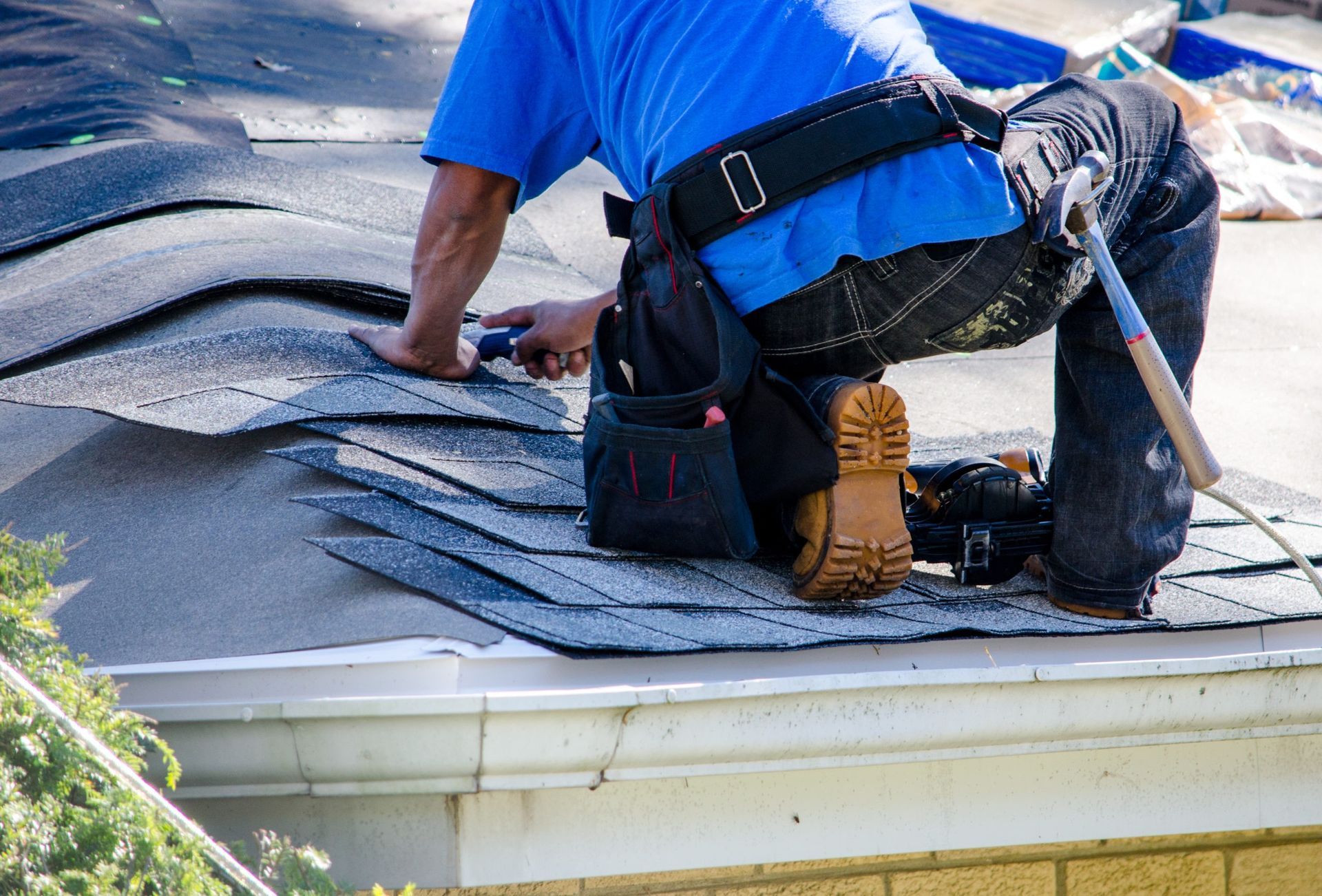 Roofer in blue shirt kneels on a shingled roof, installing dark shingles near the roof edge.