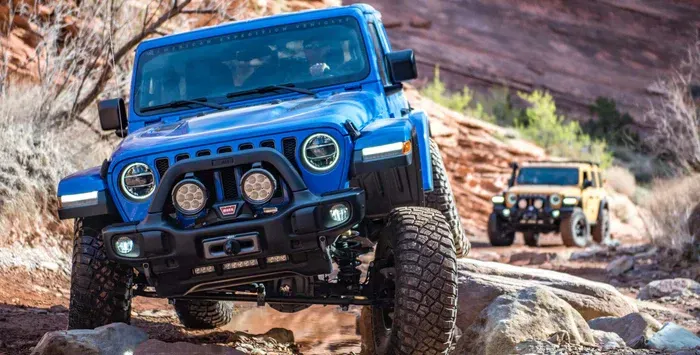 A bright blue Jeep off-roading on rocky terrain with a yellow Jeep following behind in the background.