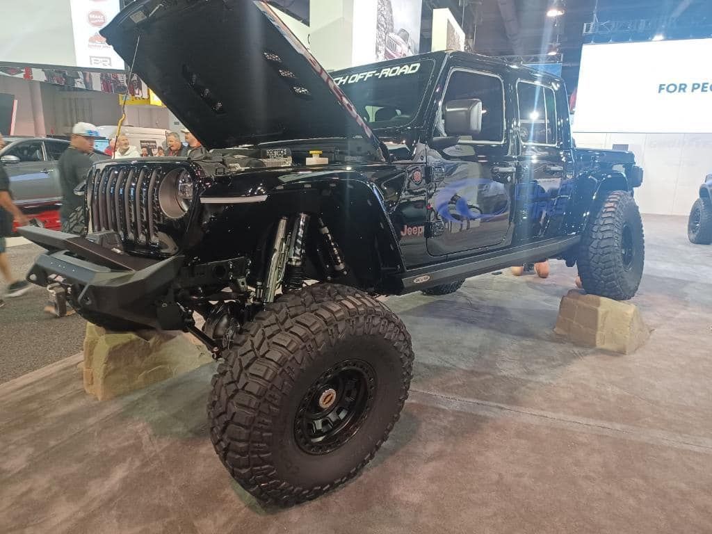 A black, highly modified Jeep with its hood open, displayed on raised blocks in an indoor exhibition hall.