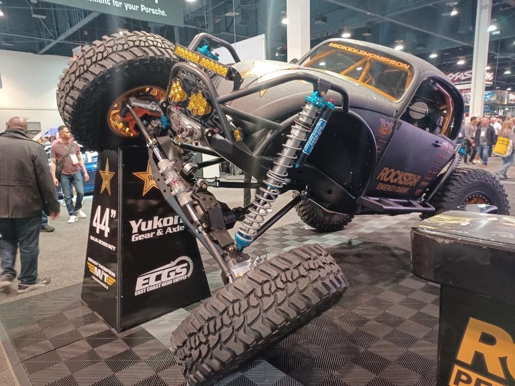 A customized off-road vehicle with large tires and exposed suspension on display at an indoor automotive trade show.